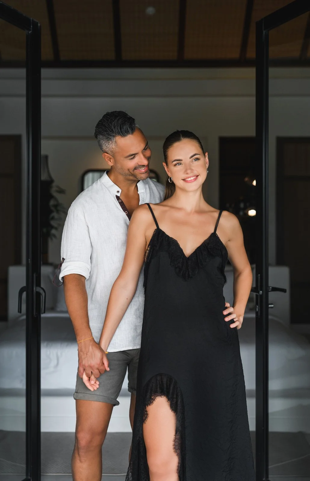 A couple standing warmly holding hands inside a modern home with a bed in the background. The woman wears a black dress with a high slit, and the man wears a white shirt with rolled-up sleeves and shorts. They appear happy and affectionate.
