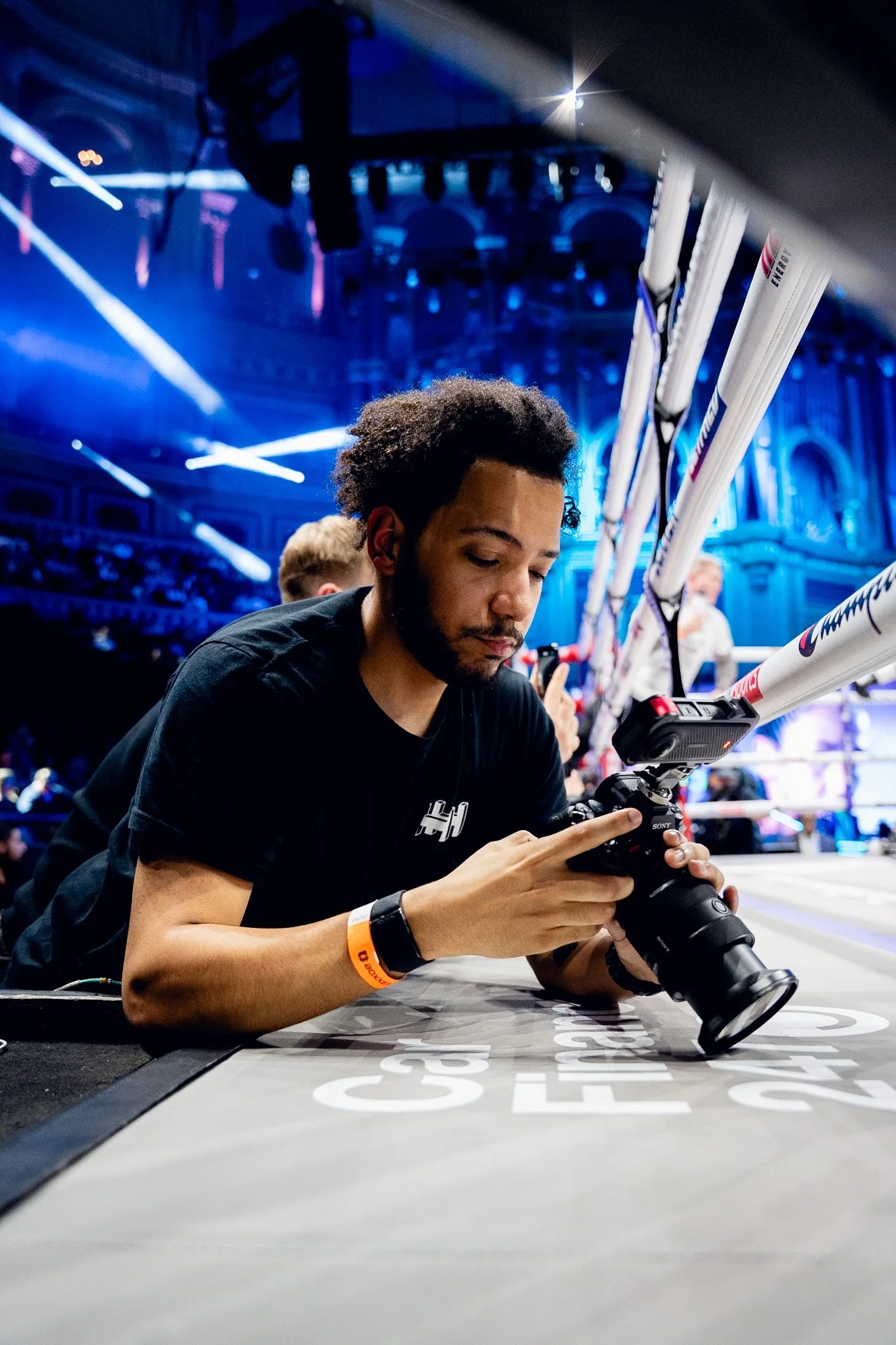 A young man with curly hair and a beard kneeling at a table, looking at a camera in his hands. He is wearing a black t-shirt with a logo on it and an orange wristband. The background features a brightly lit boxing ring with ropes, and the venue has an elaborate ceiling with arches, stage lights, and large screens.
