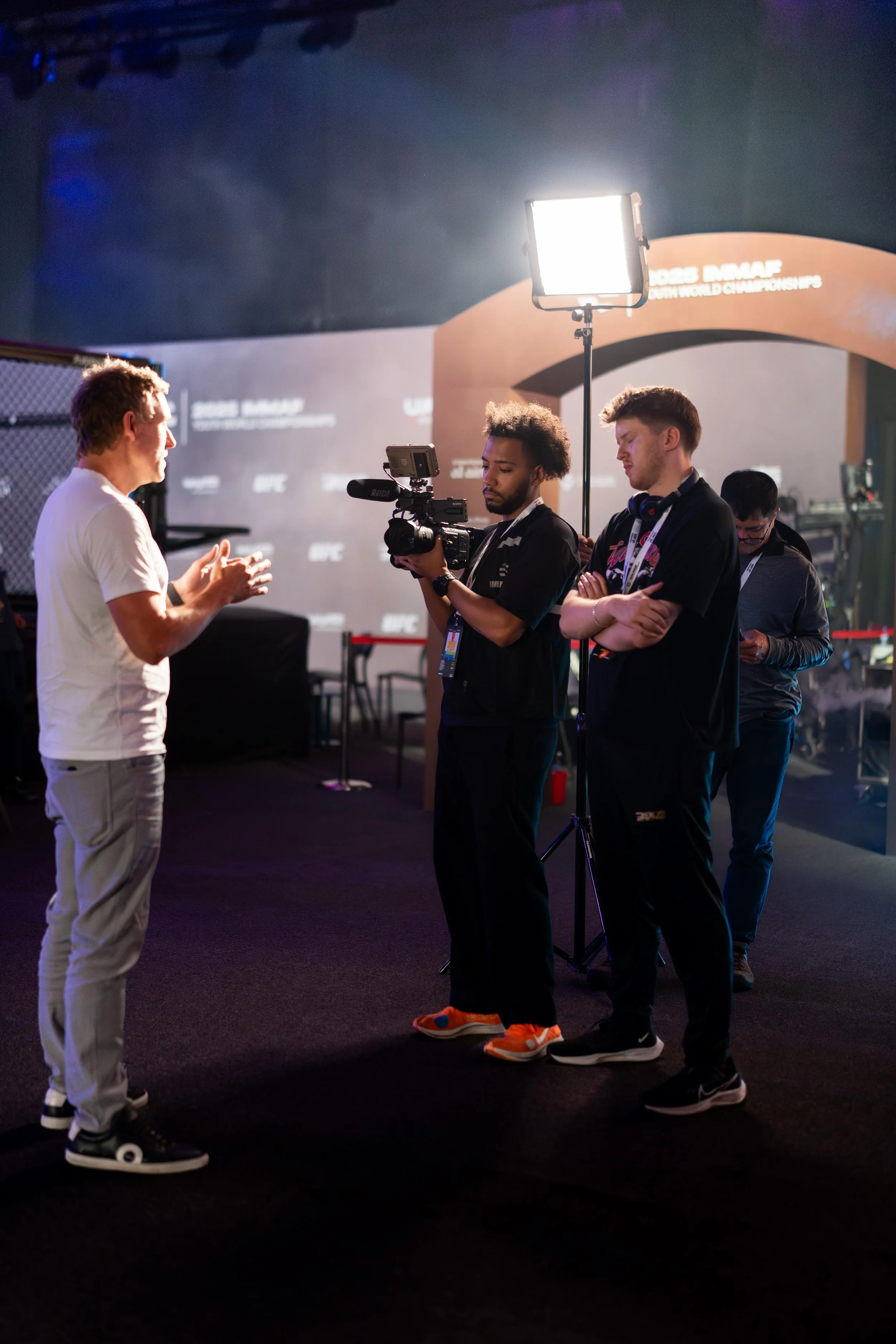 Interview or conversation taking place at a sports event, with a man in a white shirt talking to three young men in black shirts, one of whom is filming with a professional camera, and another is standing in front of a large screen with text about a sports championship.