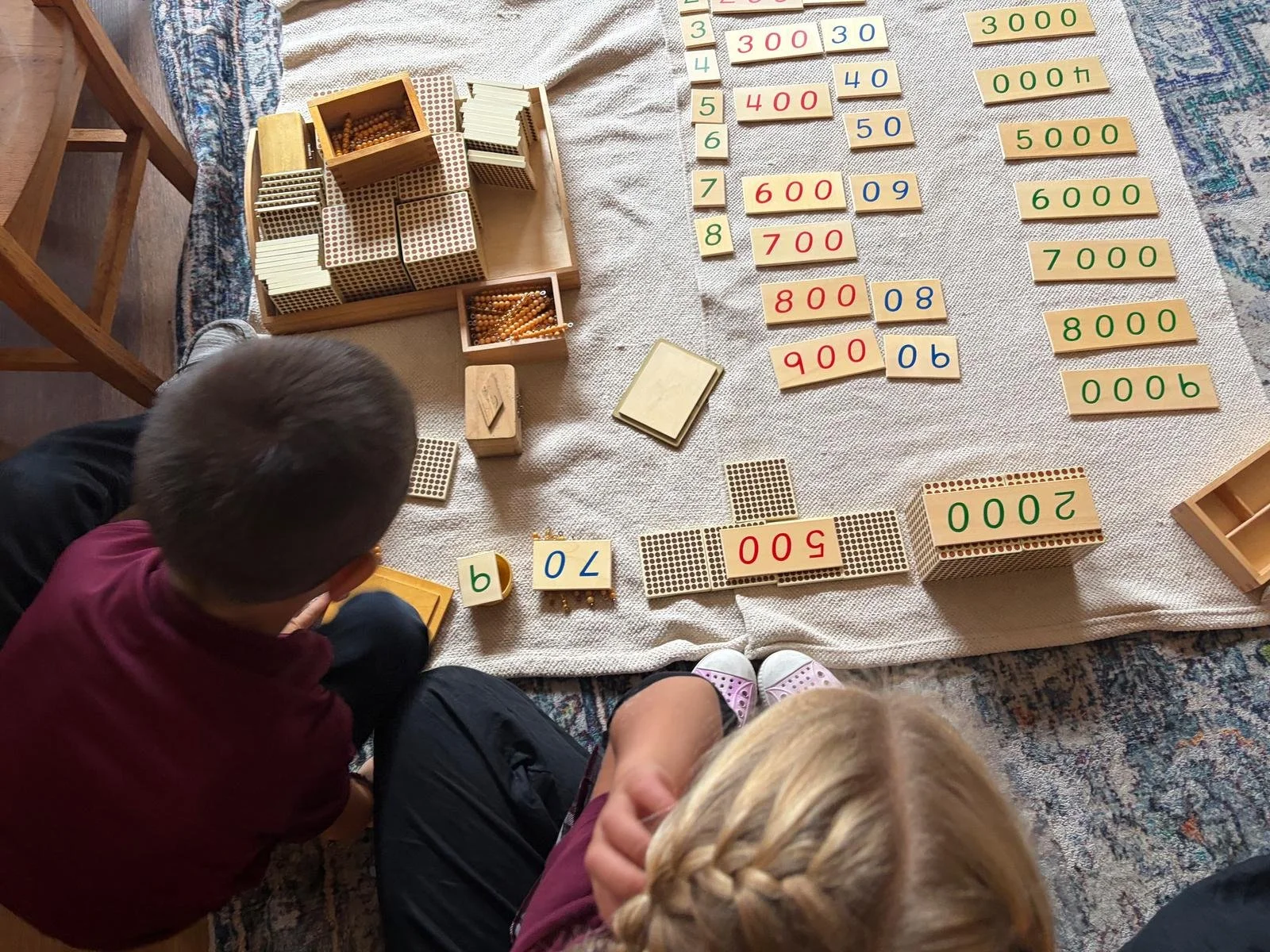 Two children and an adult sitting on the floor learning about numbers with educational toys, including number tiles, flashcards, and counting blocks, arranged on a white cloth.