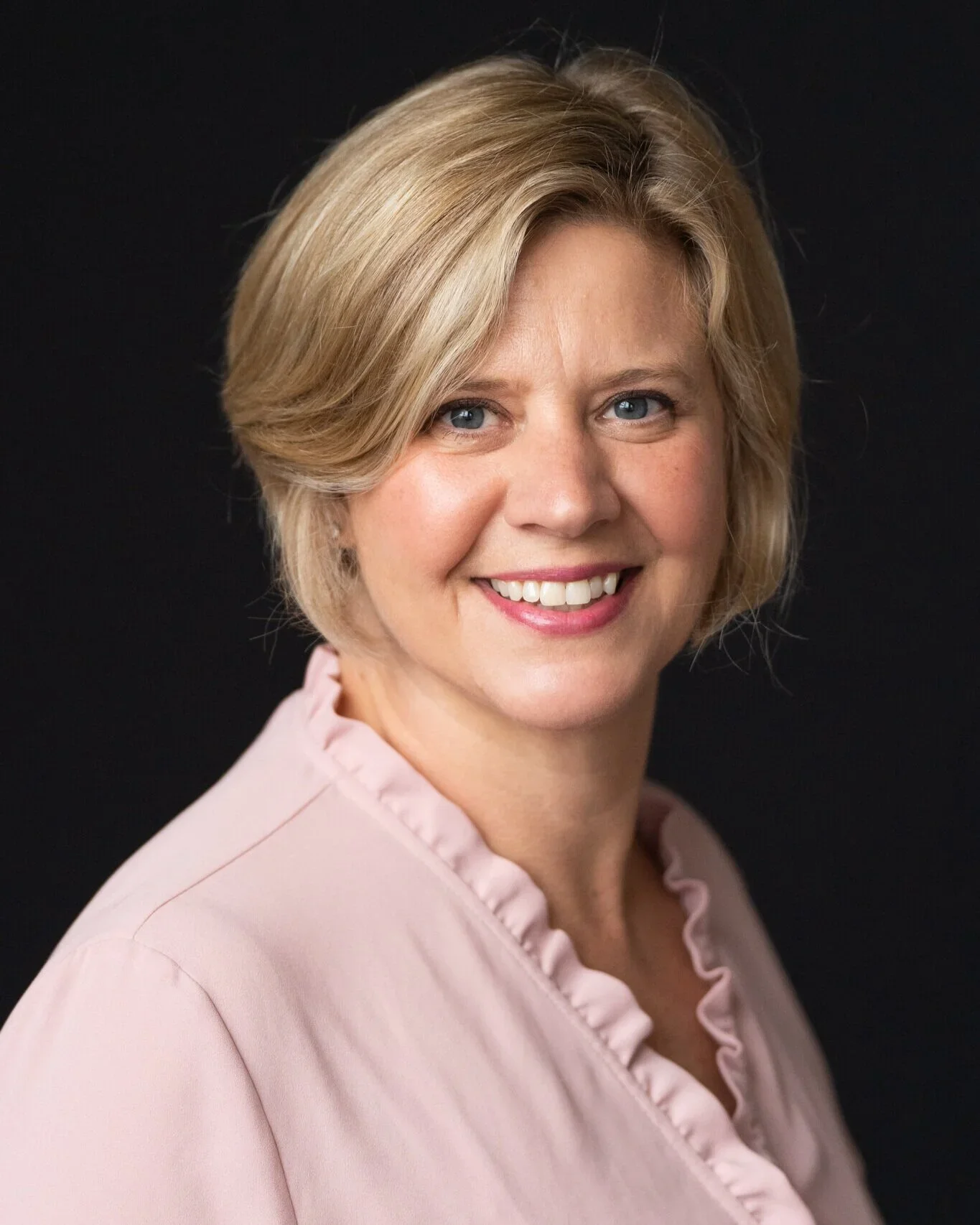 A middle-aged woman with short blonde hair, blue eyes, and a friendly smile, wearing a light pink blouse with ruffled collar, against a black background.