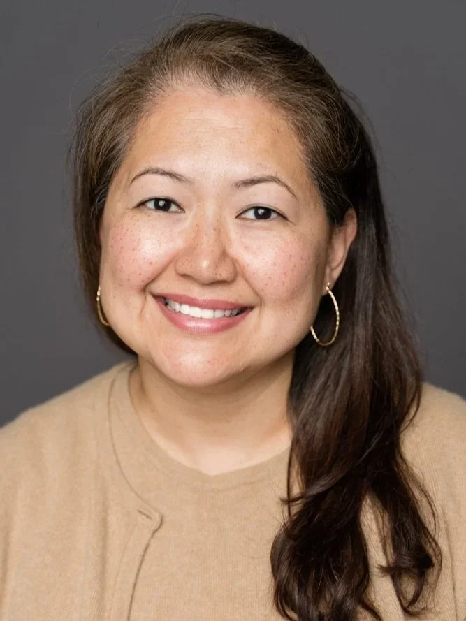 Portrait of a smiling woman with long brown hair, wearing gold hoop earrings and a beige top, against a gray background.