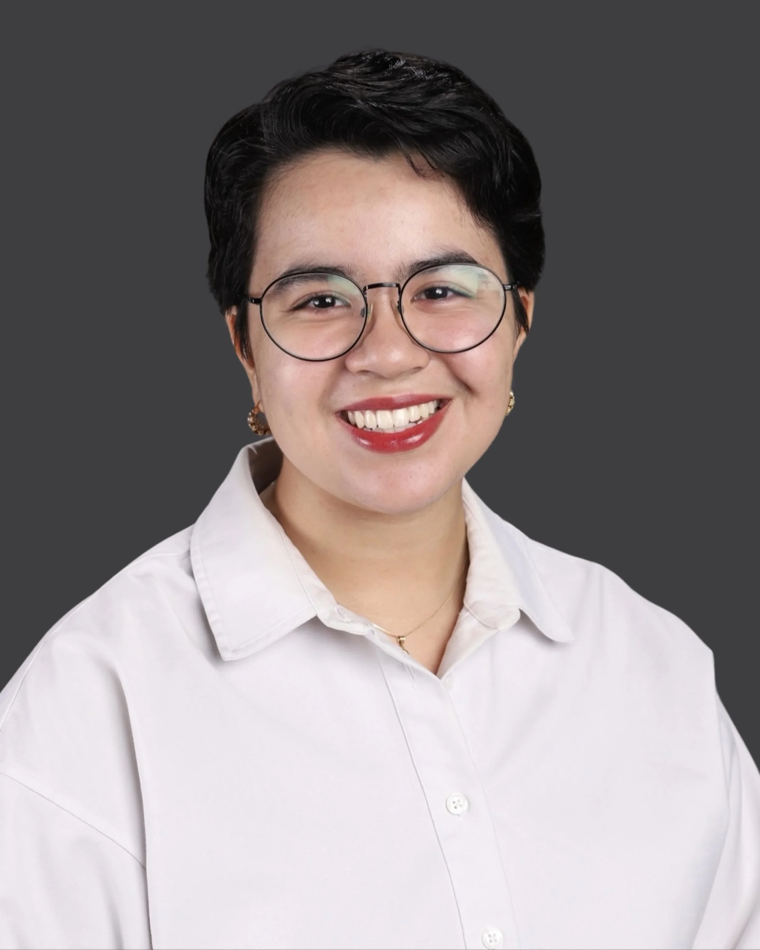 Close-up of a smiling woman with short black hair, wearing glasses and a white button-up shirt, against a dark grey background.