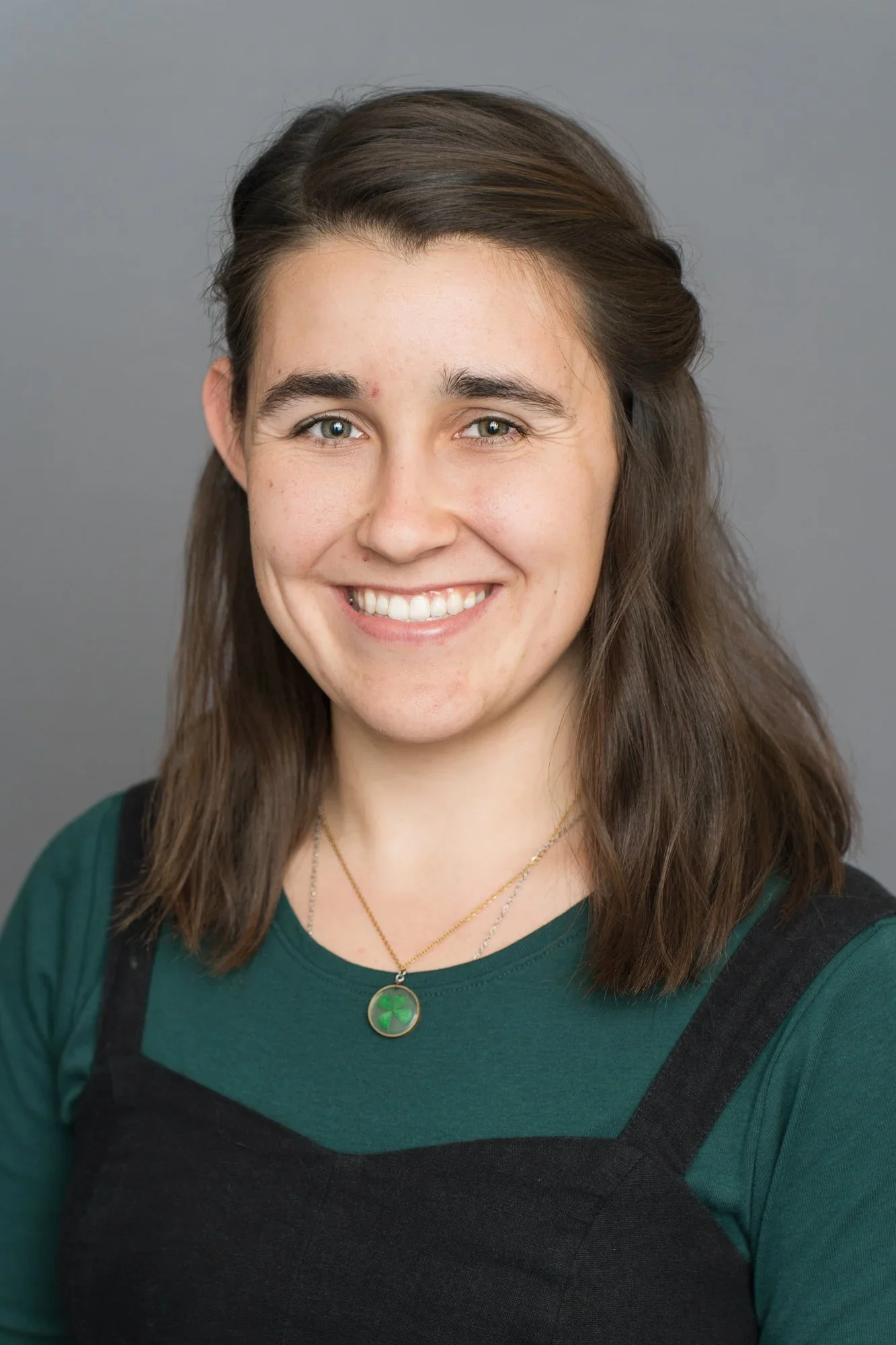 A young woman with long brown hair, light skin, and blue eyes smiling, wearing a green top with black straps, and a necklace with a green pendant against a plain grey background.
