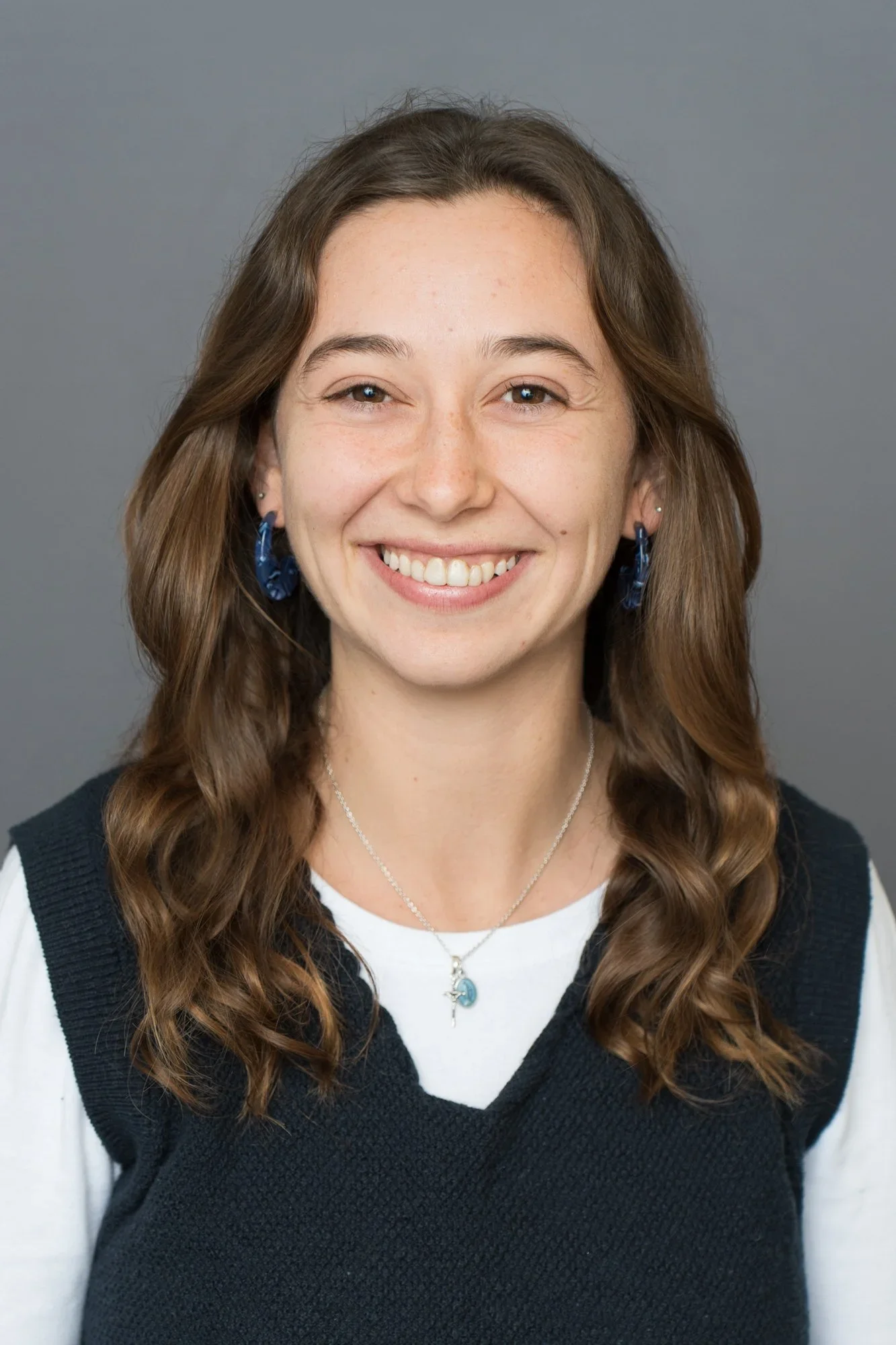 A young woman with long, wavy brown hair and fair skin, smiling, wearing blue earrings and necklaces against a gray background.