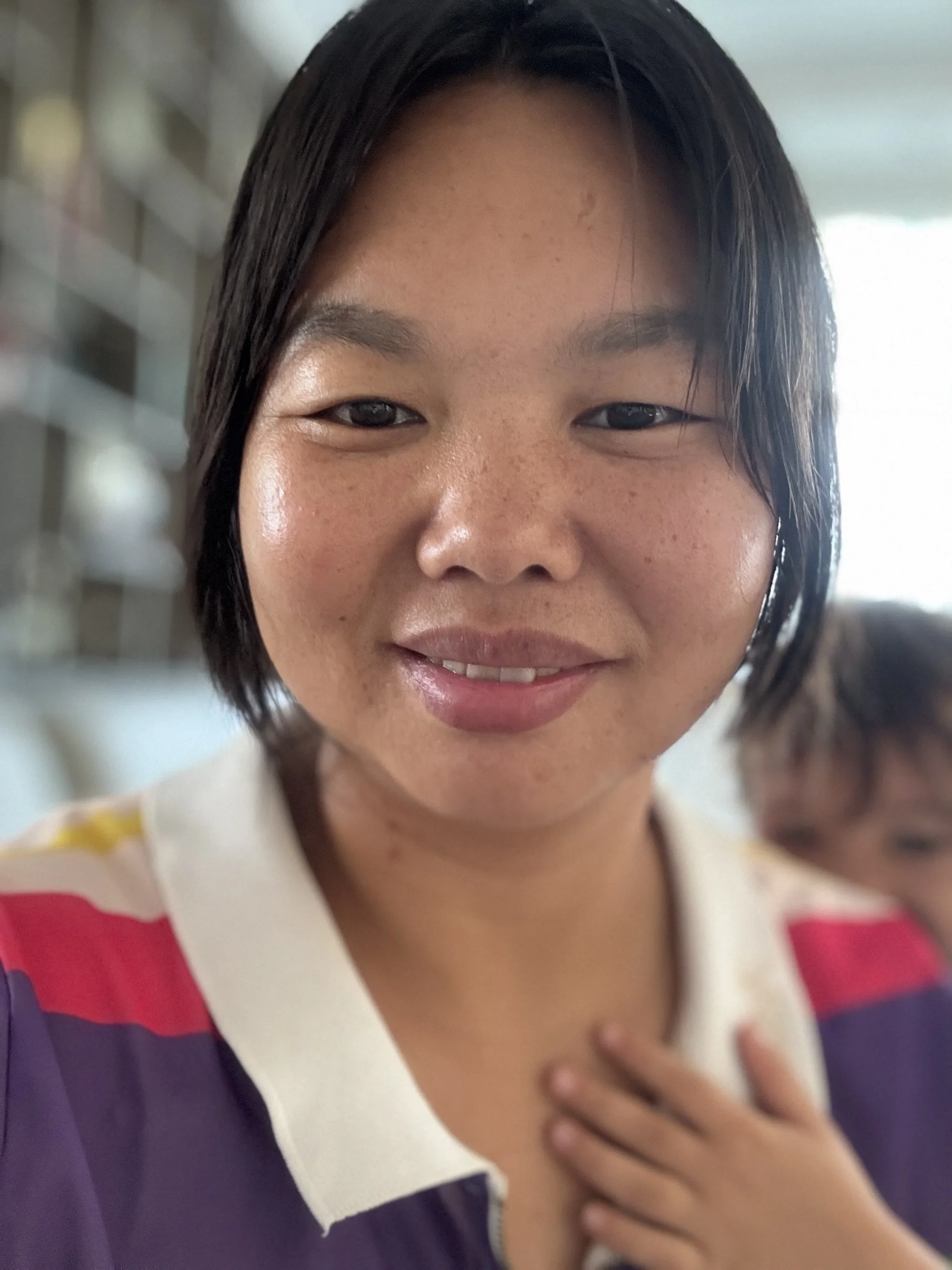 Close-up of a smiling young Asian girl with short black hair, wearing a colorful shirt, with her hand on her chest, in an indoor setting.