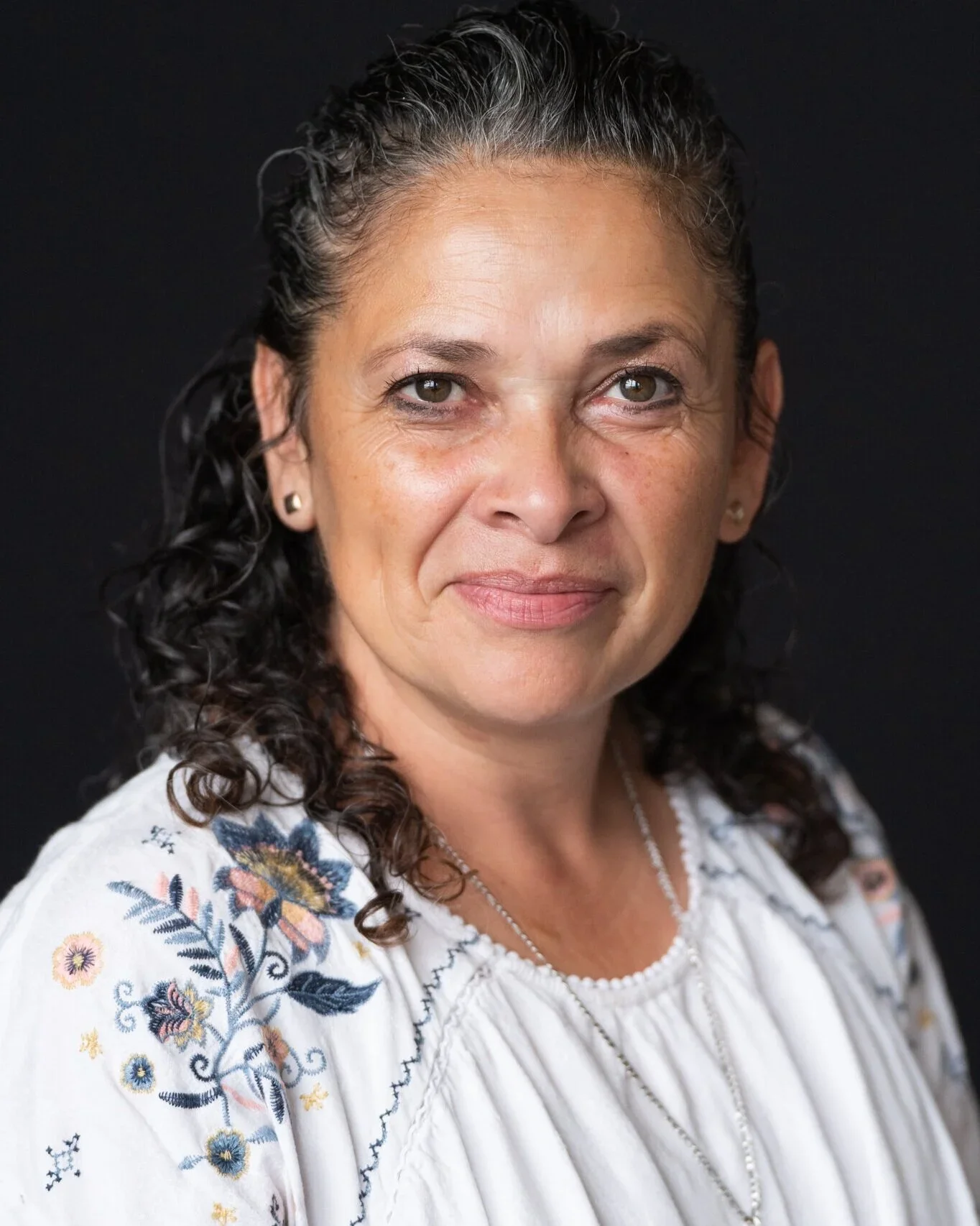 A middle-aged woman with dark curly hair, light skin, and brown eyes, wearing a white embroidered blouse with floral patterns and jewelry, standing against a black background.