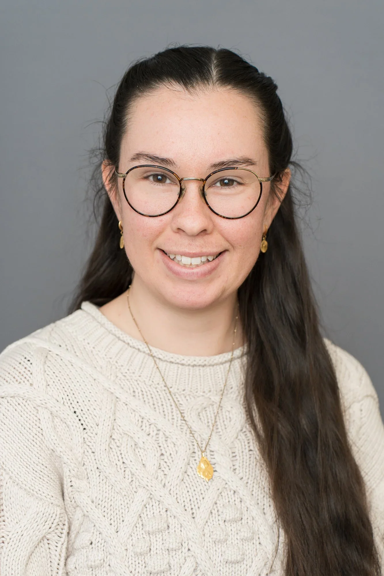 A woman with long dark hair, round glasses, and gold earrings, smiling while wearing a white knit sweater and a gold necklace, standing against a gray background.