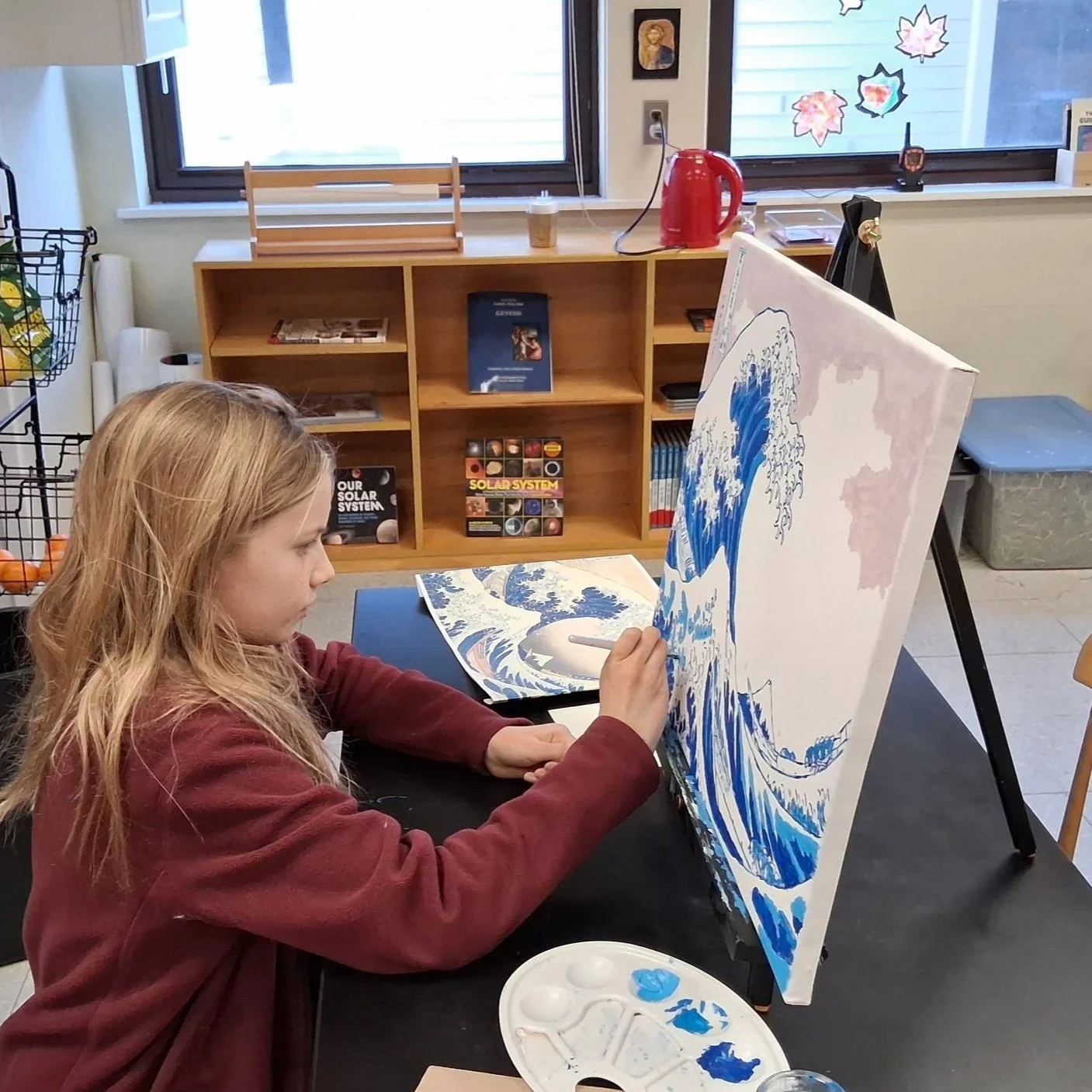 A young girl concentrating while painting a replica of 'The Great Wave off Kanagawa' on a canvas in an art classroom. She is seated at a table with an artist's palette, in a room with shelves of books about the solar system and decorated with autumn 