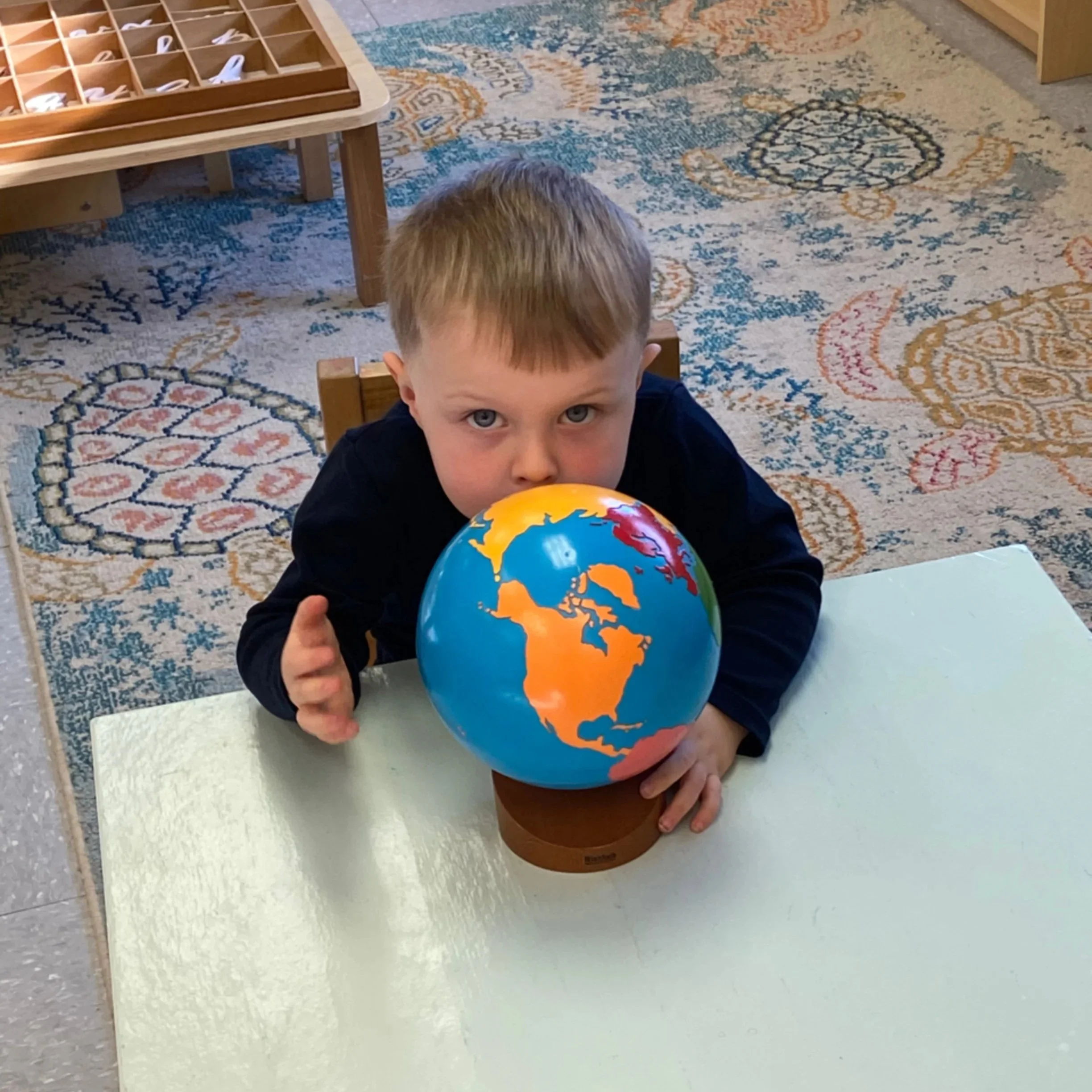 A young boy with blond hair and blue eyes playing with a colorful globe in a classroom with a patterned rug.