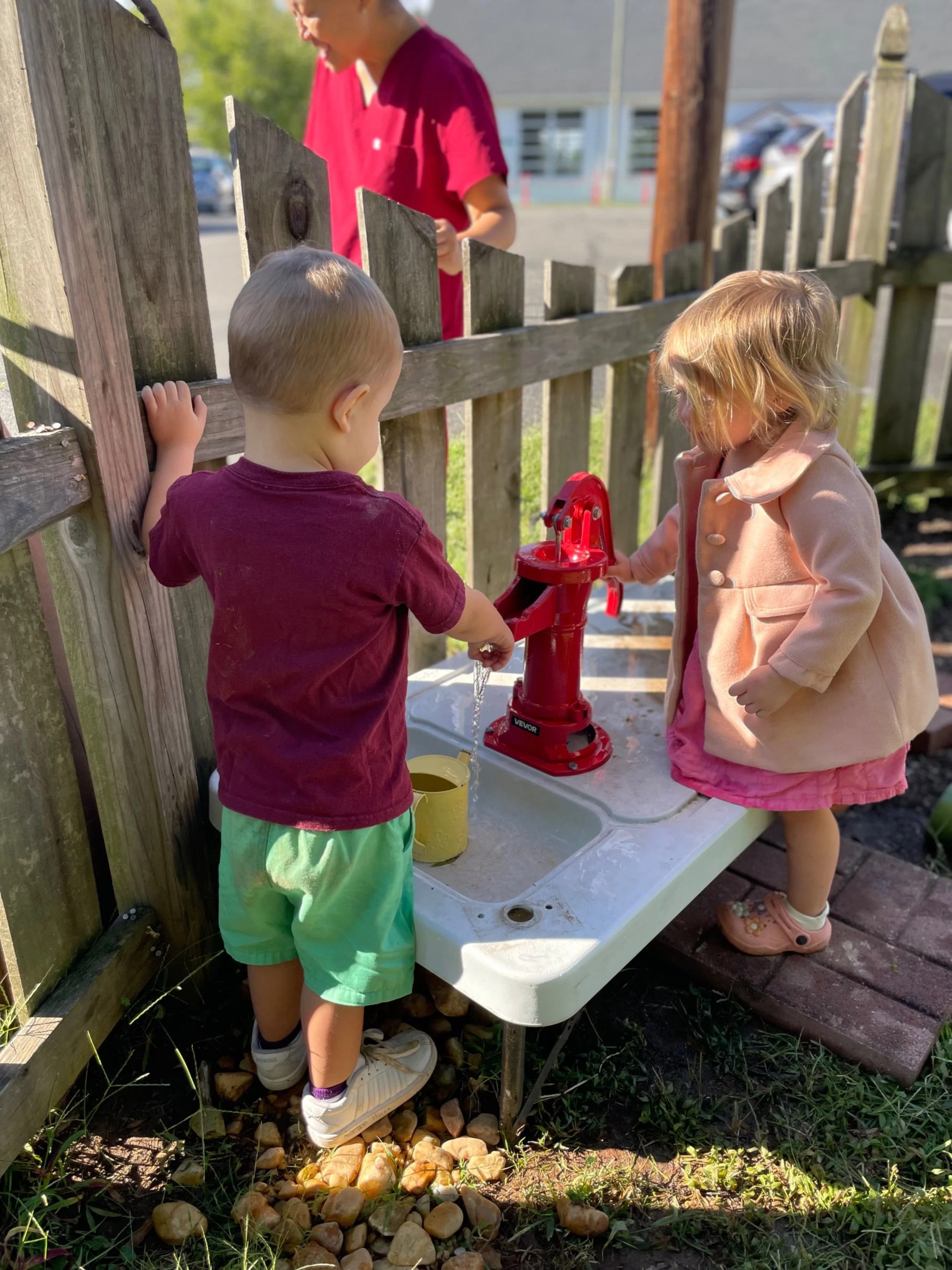 Two young children, a boy with blonde hair in a maroon shirt and light green shorts, and a girl with blonde curly hair in a pink jacket, are playing at a water pump outdoors. The boy is pressing the handle of the red water pump, and the girl is looking at it, both standing by a makeshift outdoor sink. Another child in a red shirt is partially visible behind a wooden fence in the background, along with parked cars and a building.