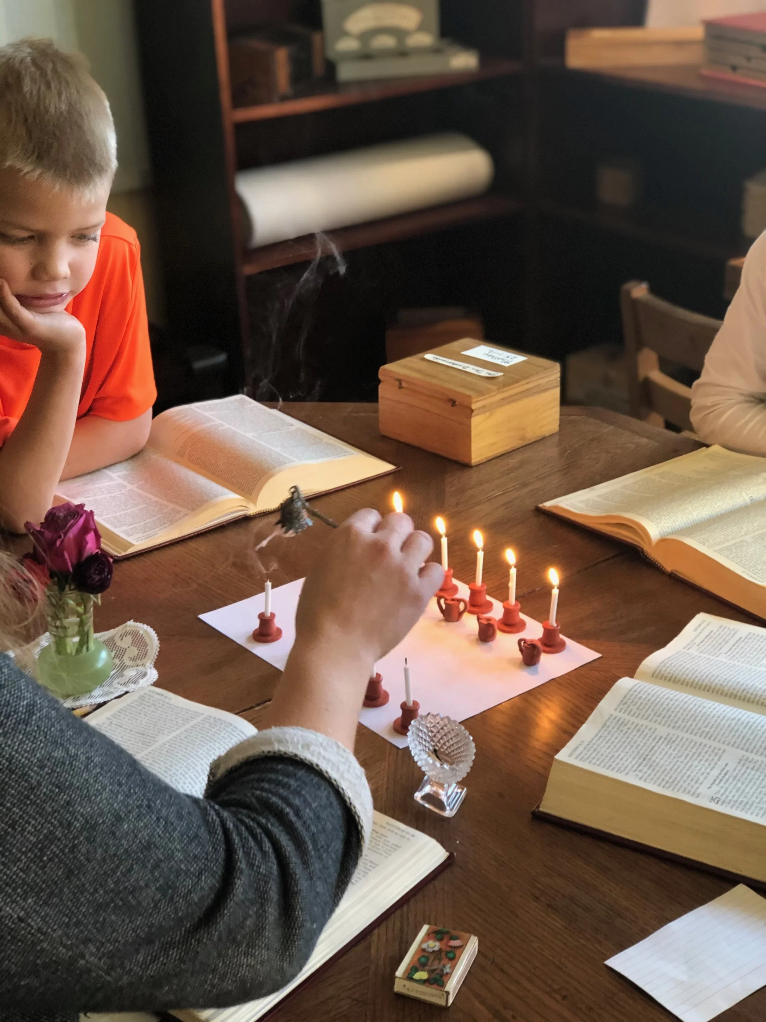 Children gathered around a table with open books, playing chess with lit candles. There are flowers and various items on the table, and bookshelves in the background.
