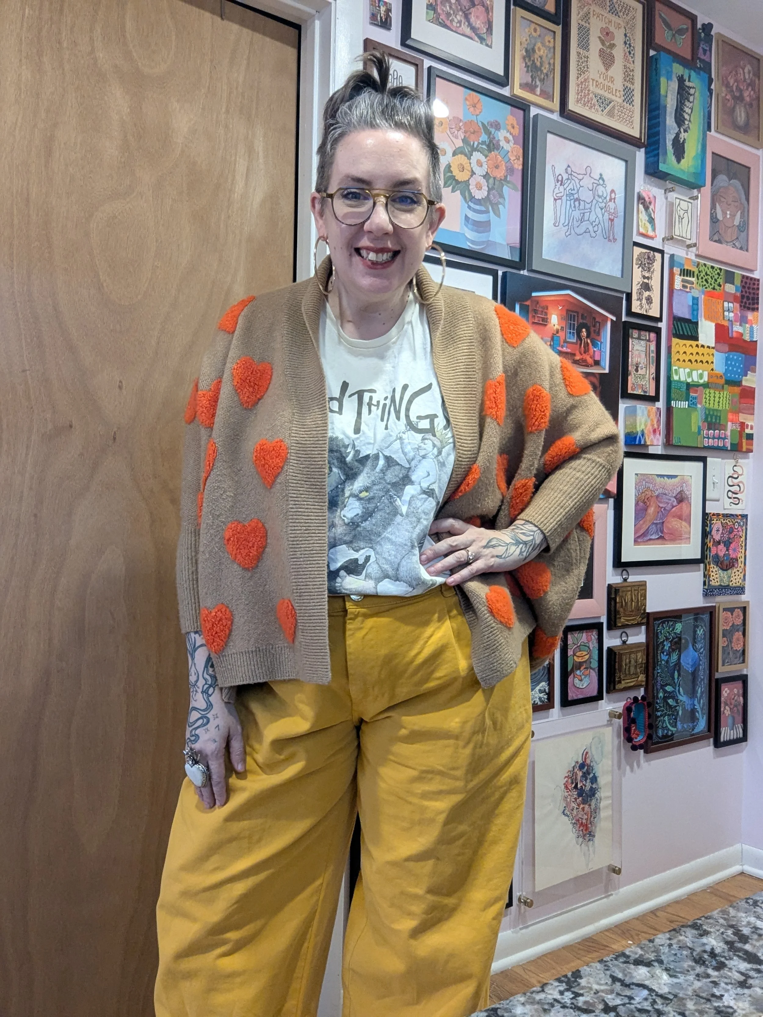 A woman with glasses, hoop earrings, and tattoos on her hand and arm, smiling and standing with her hand on her hip in front of a wall decorated with colorful framed artwork. She is wearing a beige cardigan with orange hearts and yellow pants.