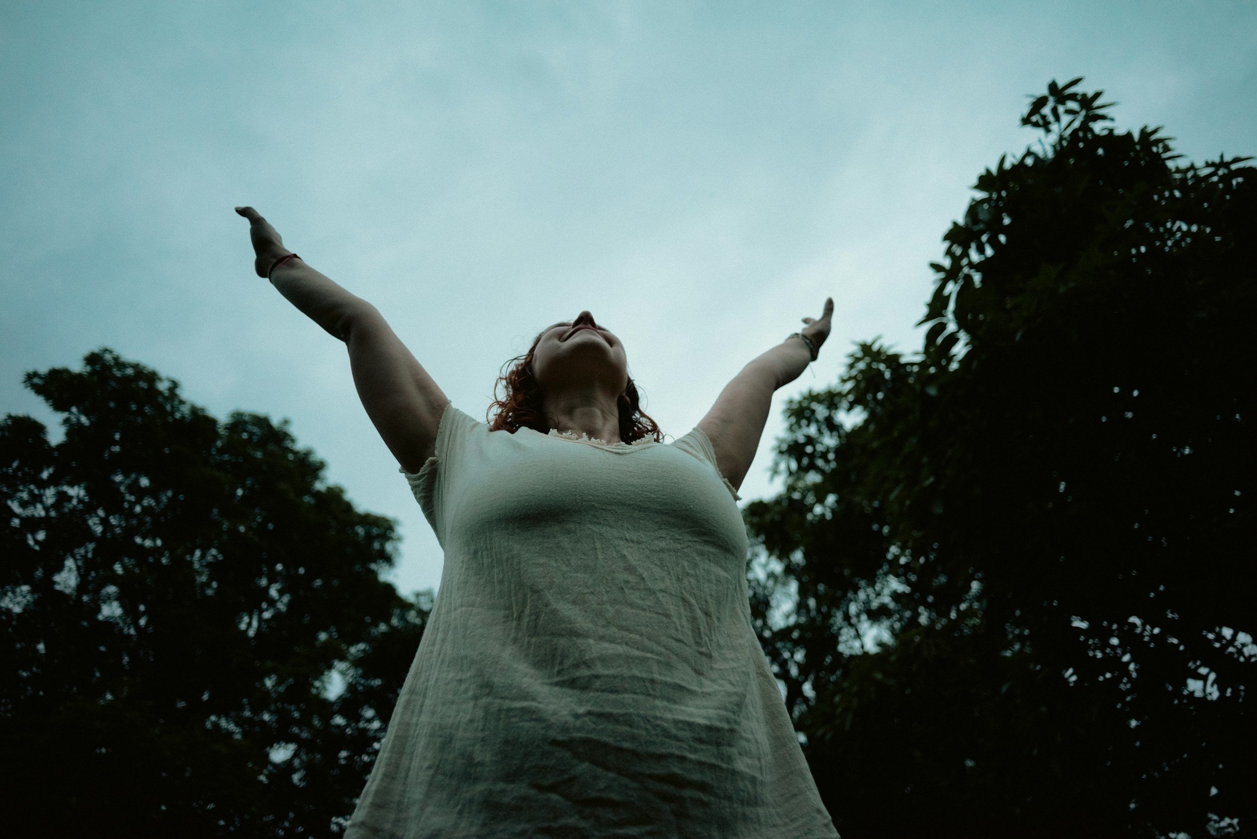 A woman with red hair wearing a white dress standing outdoors with her arms raised towards the sky, surrounded by trees.