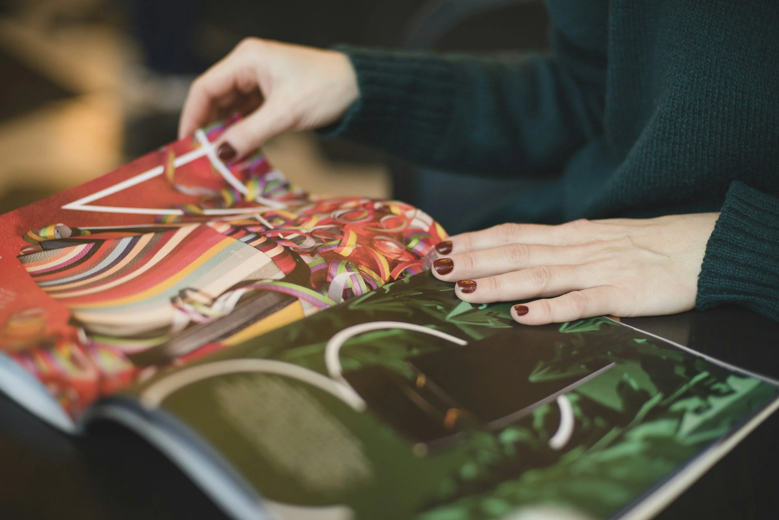 Close-up of hands with dark nail polish wrapping colorful ribbons on a gift, with a patterned gift bag nearby.