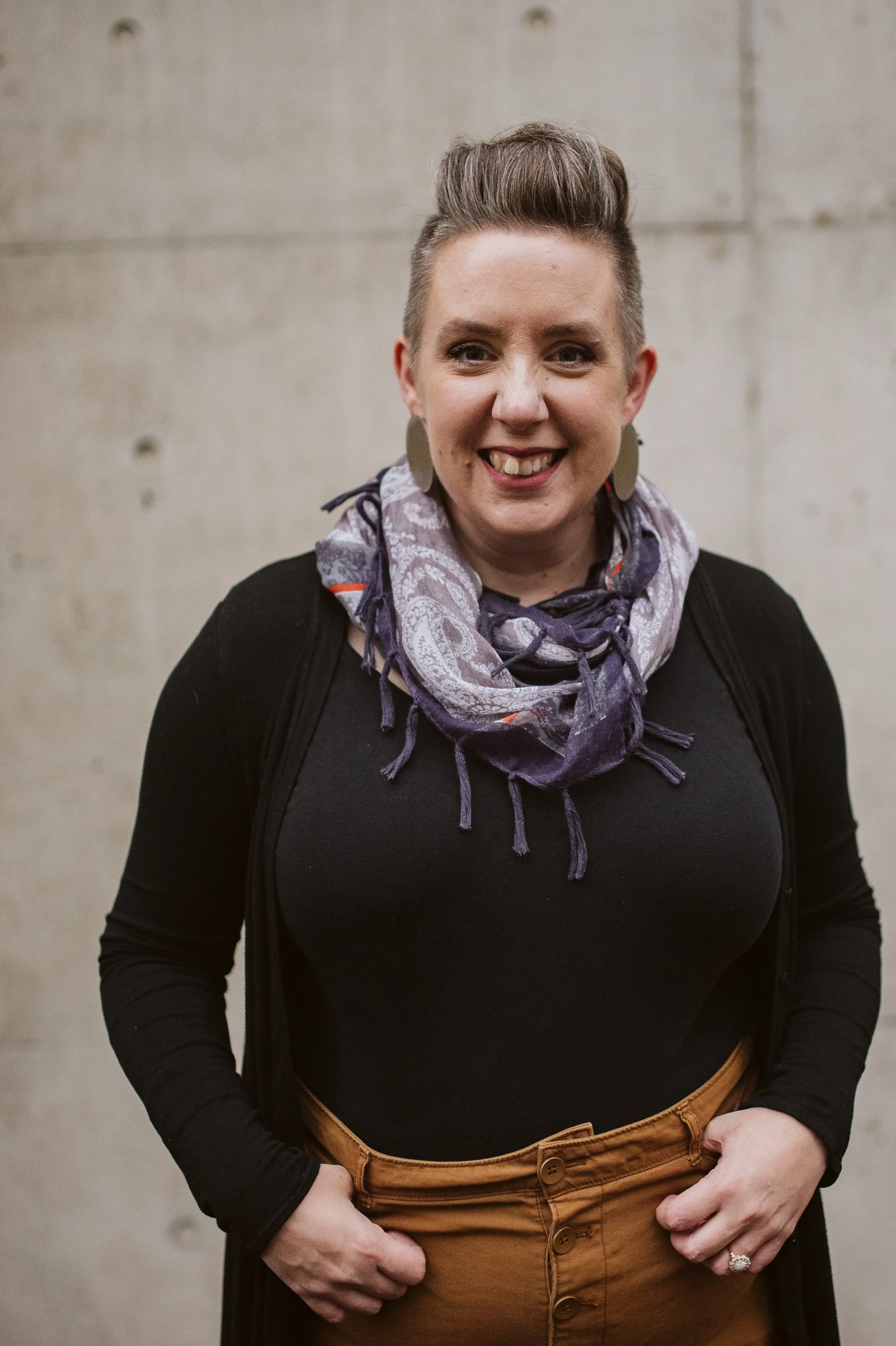 A woman smiling, wearing a dark long-sleeve shirt, patterned scarf, tan high-waisted pants, and earrings, standing against a neutral background.
