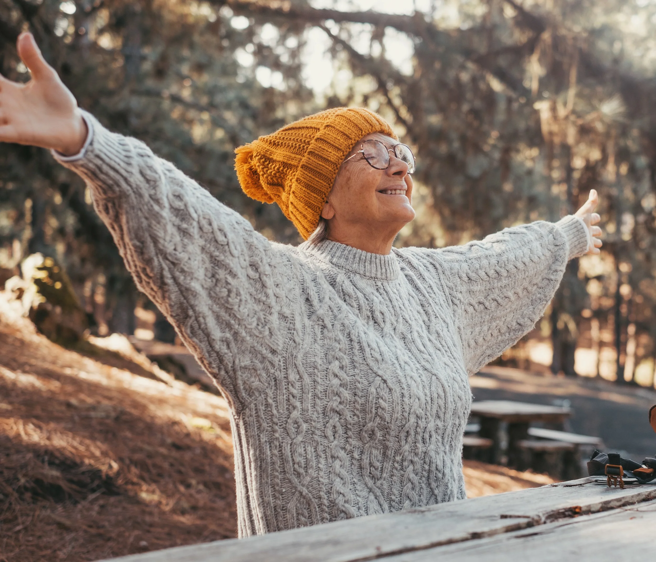 An elderly woman wearing a yellow knit hat and glasses with arms outstretched outdoors in a park with trees and benches.