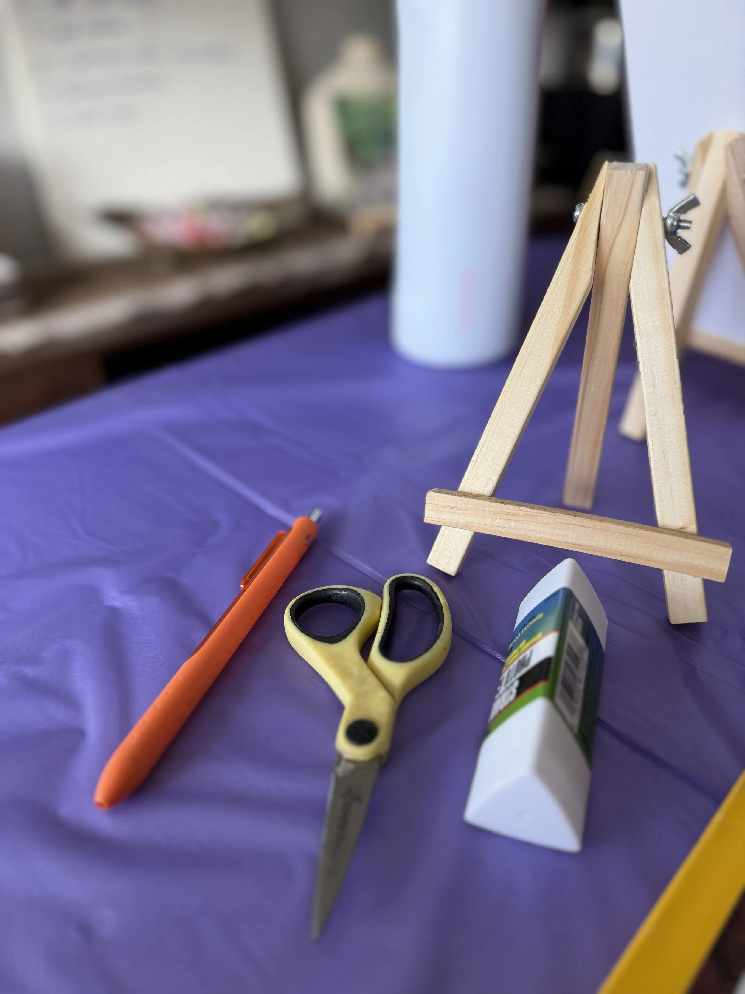 A workspace with a wooden easel, a pair of scissors, a marker, a glue stick, and a smartphone on a purple tablecloth.
