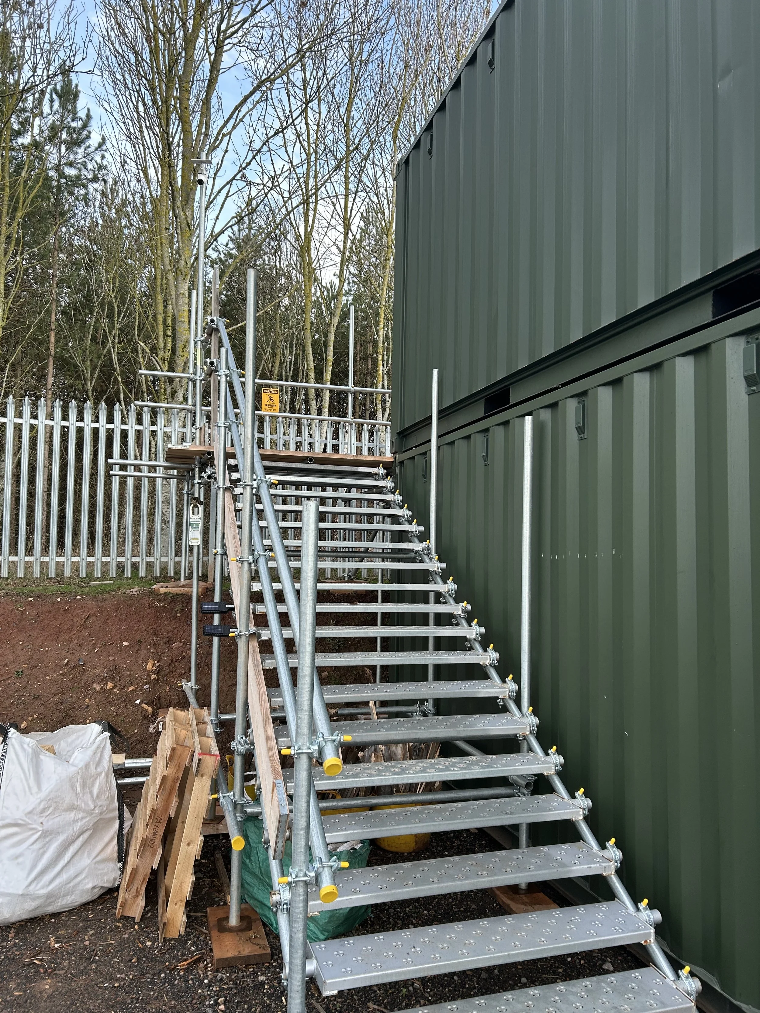 Metal scaffolding set up beside a green container with a ladder and construction materials at an outdoor site with trees in the background.
