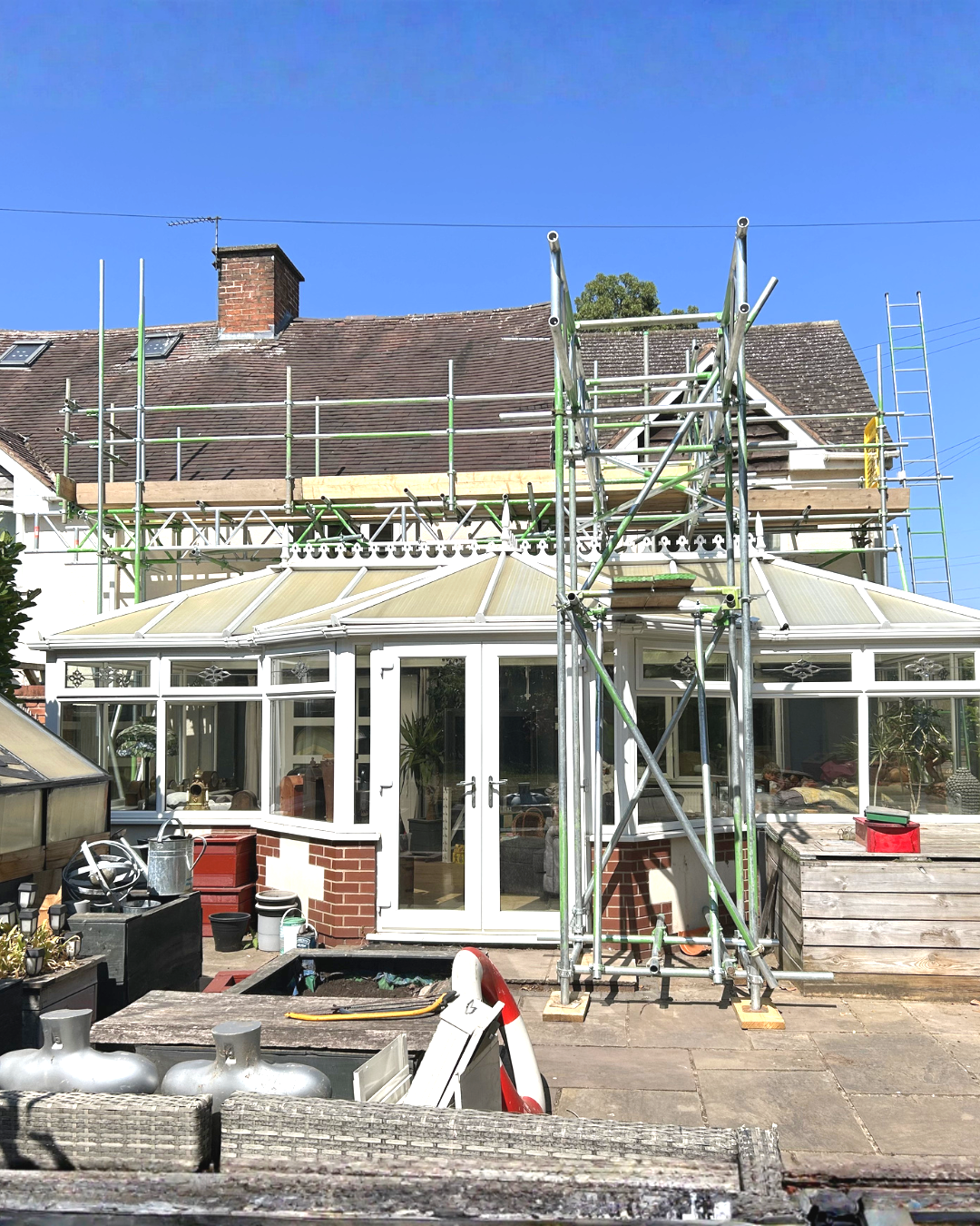 Scaffolding set up around a house undergoing renovations, including a conservatory with large windows, in bright daylight with a clear blue sky.