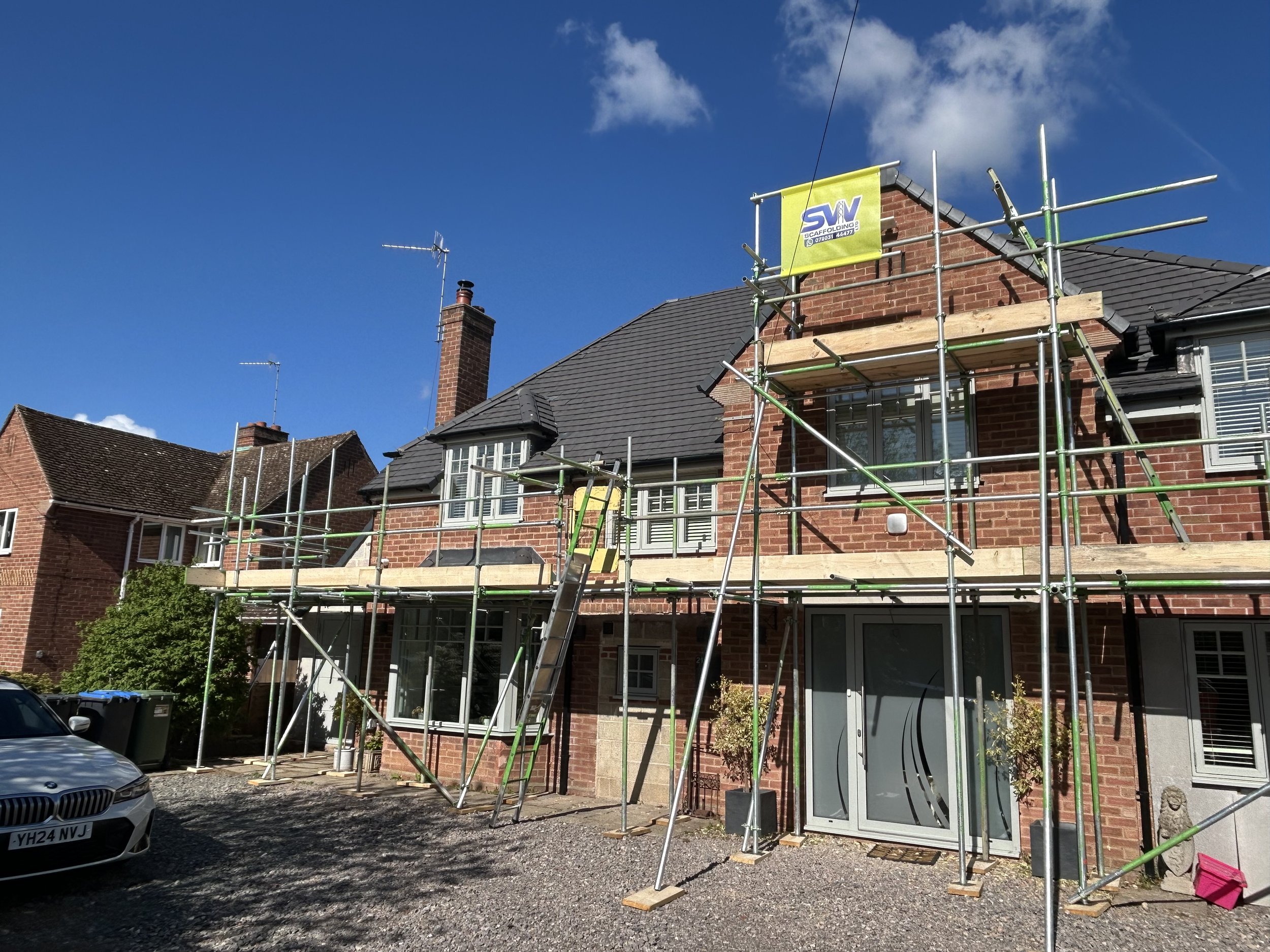 A brick house with scaffolding set up around the chimney and roof, indicating ongoing chimney or roof work on a sunny day with blue sky and some clouds. Scaffolding for homes near me, Worcestershire, Warwickshire and South Birmingham areas.
