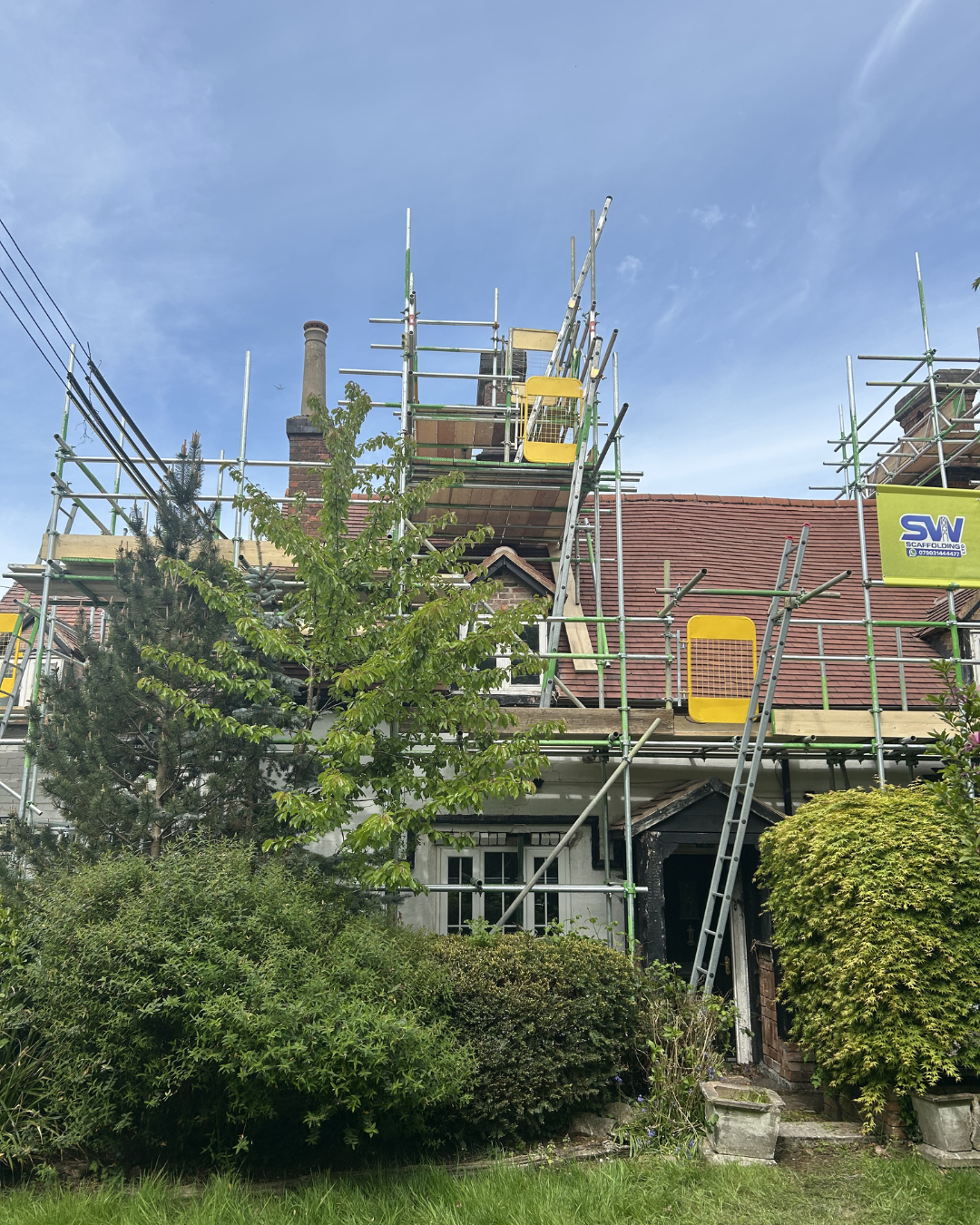 A house under renovation with scaffolding surrounding the roof. The house has a black front door, several bushes in the front yard, and a red tiled roof. The sky is blue with a few clouds.