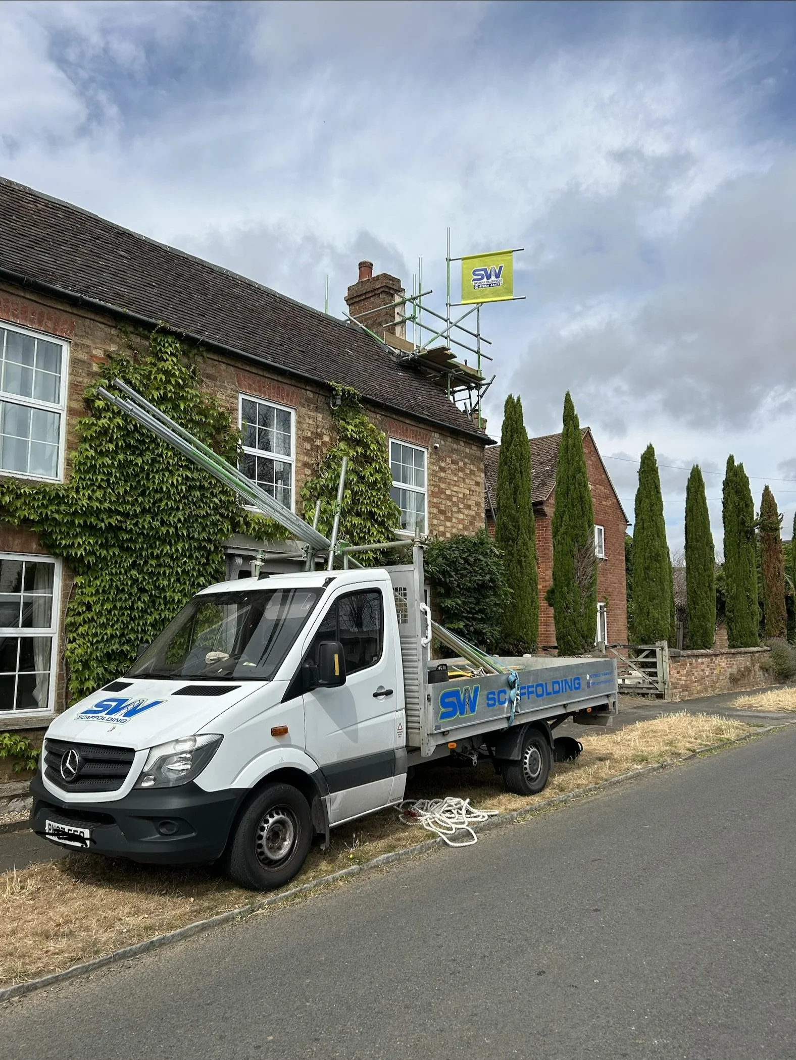 A white Mercedes-Benz utility truck with scaffolding equipment parked in front of a brick house with ivy-covered walls and tall, narrow trees.