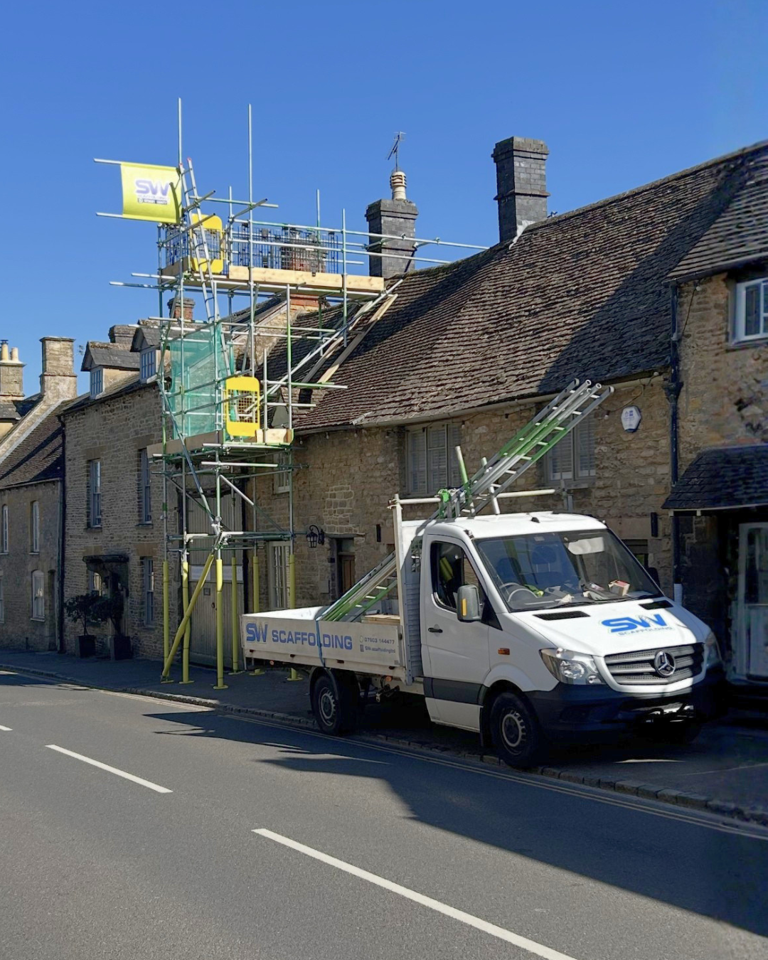 A truck with scaffolding equipment parked in front of a stone building with a sloped roof and chimneys, under a clear blue sky.