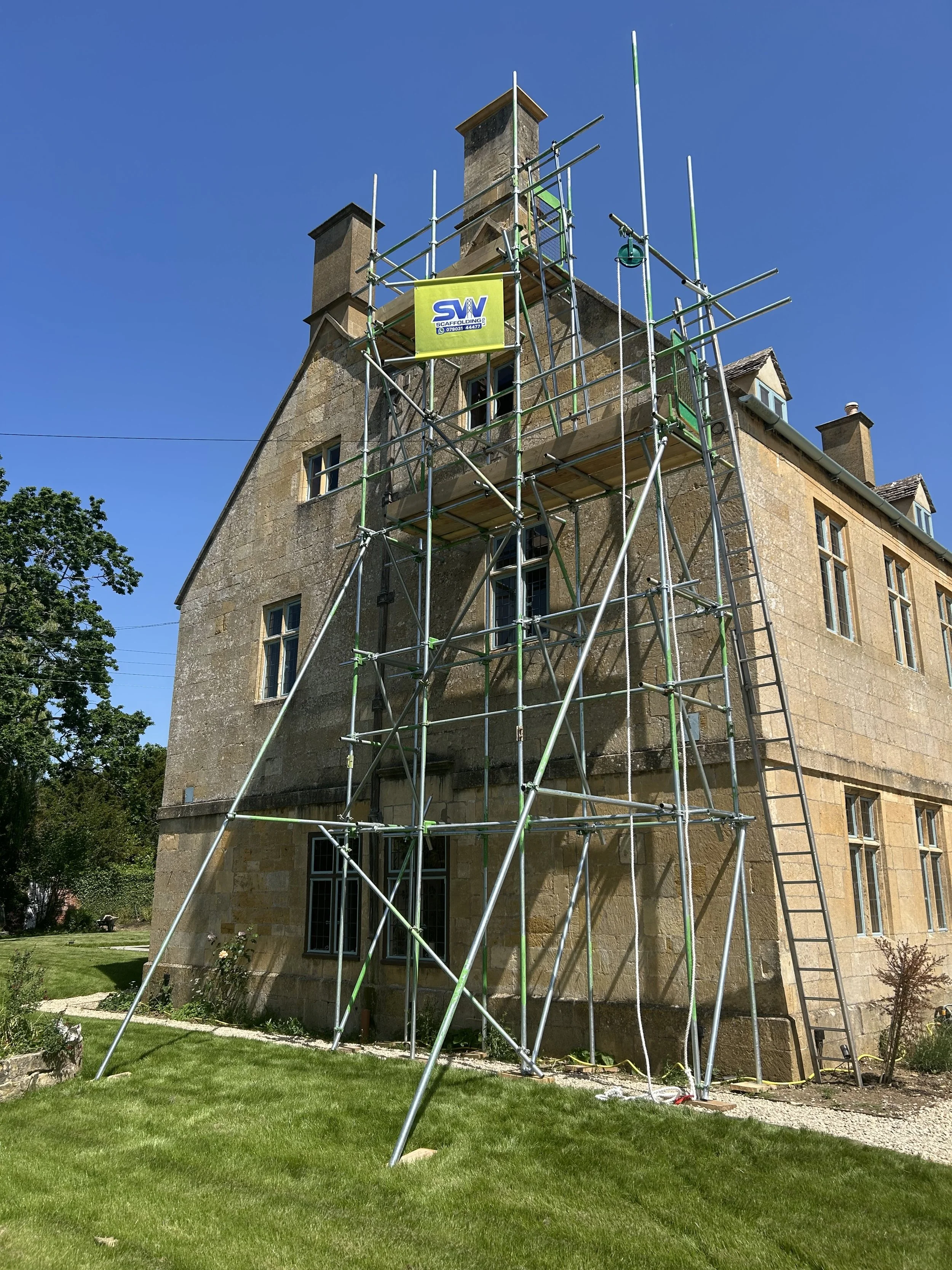 A large stone building with several windows and a chimney. Scaffolding surrounds part of the building, with a yellow sign that reads 'SIV Scaffolding.' The sky is clear and blue, and there is a well-maintained green lawn in the foreground.