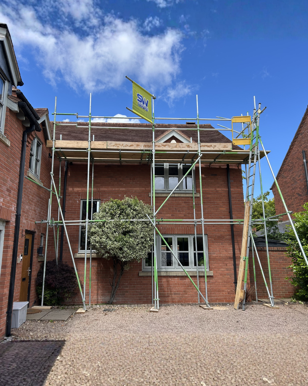 Scaffolding set up around a red brick house with a partially damaged roof, under a partly cloudy blue sky.