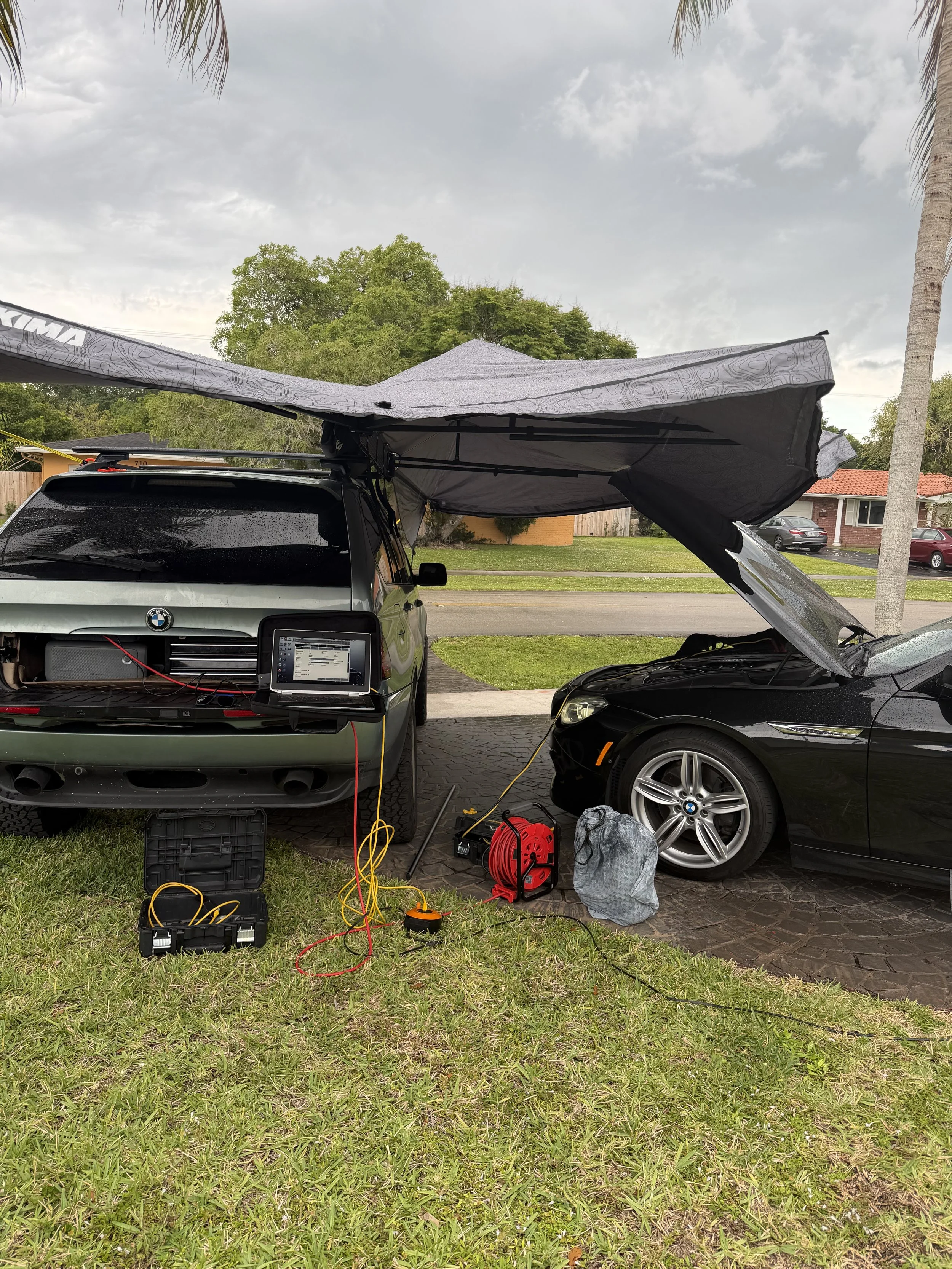 Two cars parked in a driveway with rooftop tents and equipment, with grassy yard and residential houses in the background, under cloudy sky.