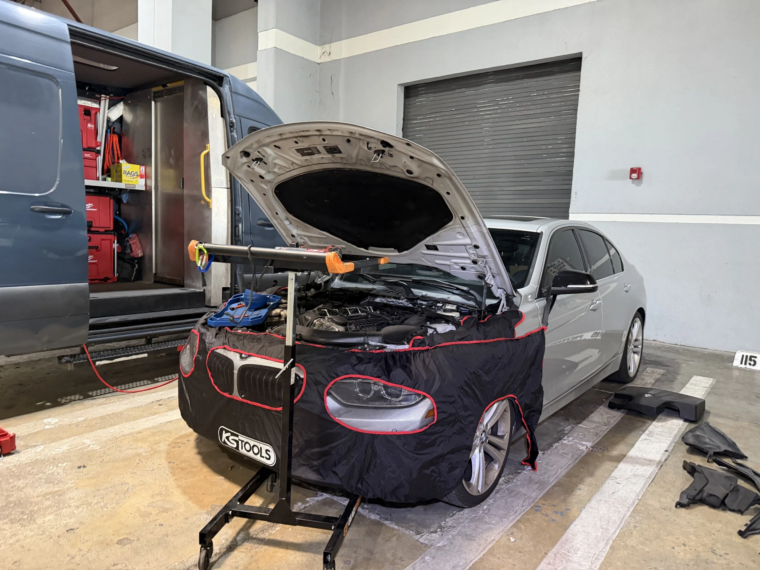 A silver car with its hood open in a parking garage, with tools and equipment around it indicating an auto repair or maintenance work. A black tool bag with red trim is in front of the car, and a roll-up garage door is in thebackground.