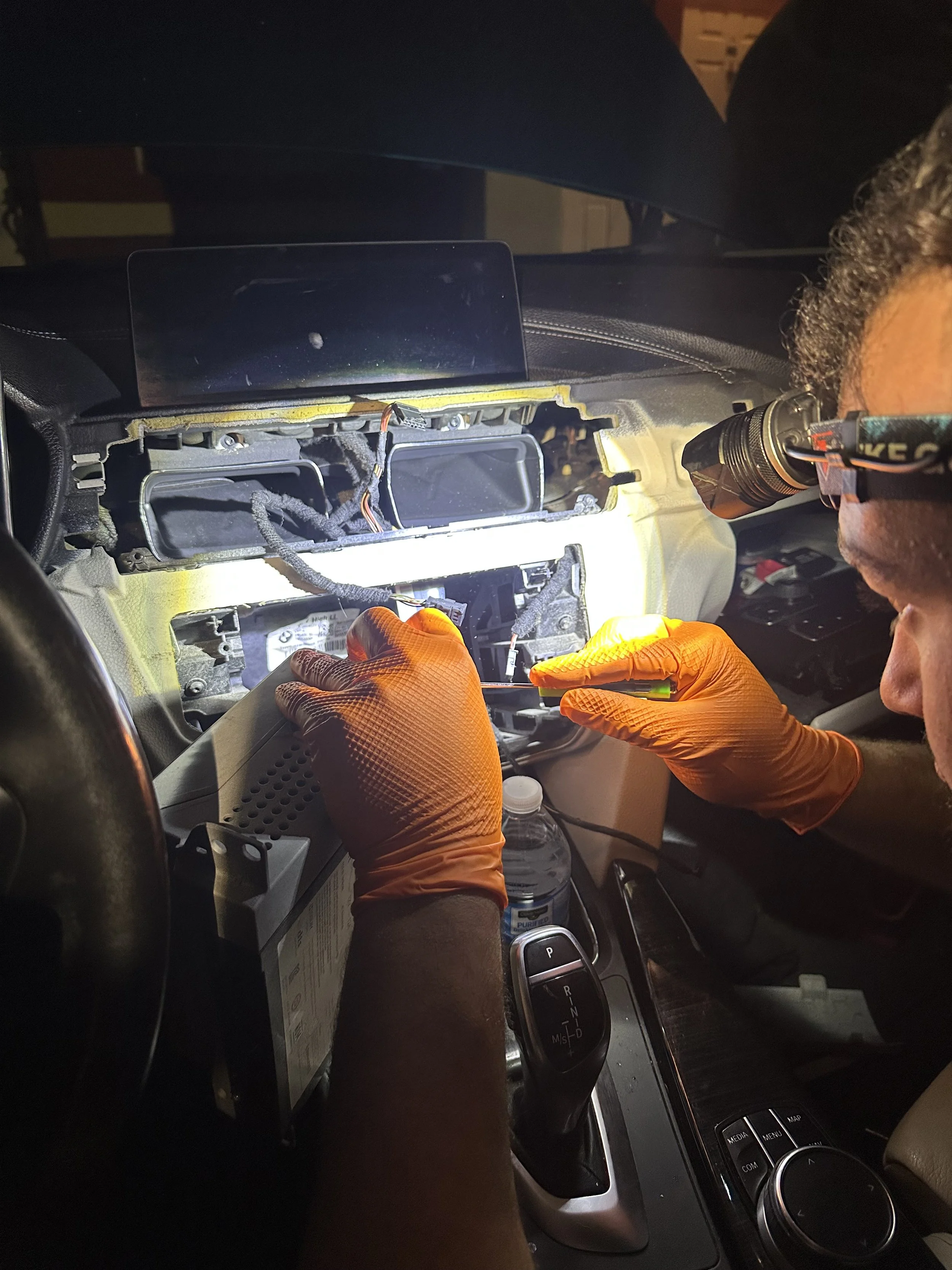 A person working inside a car, using a flashlight to inspect or repair the dashboard area. The dashboard is disassembled, exposing internal components and wiring.