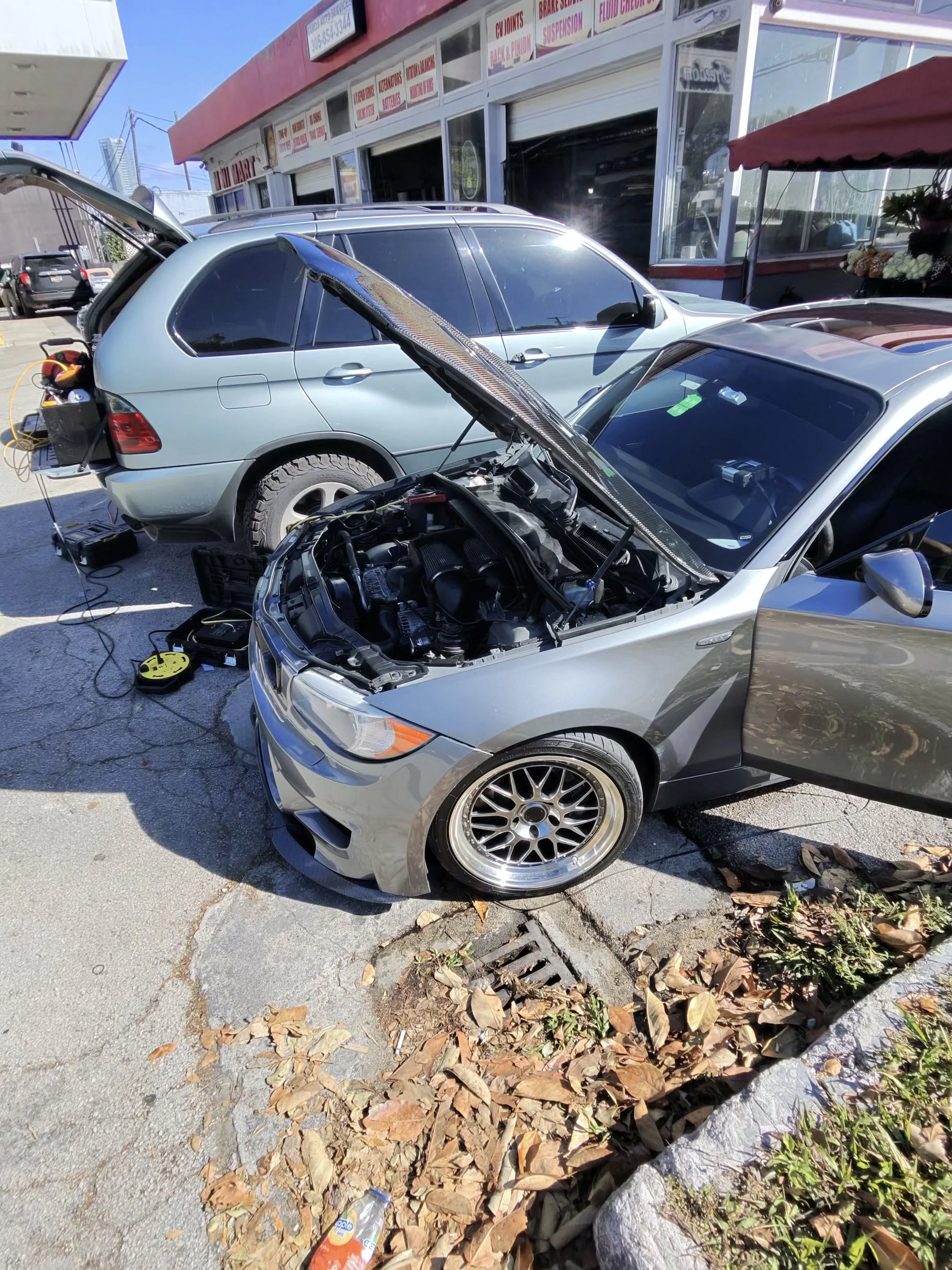 A silver car with its hood open parked outside a shop, with car maintenance tools and a battery charger nearby. Fallen leaves and trash are on the sidewalk.