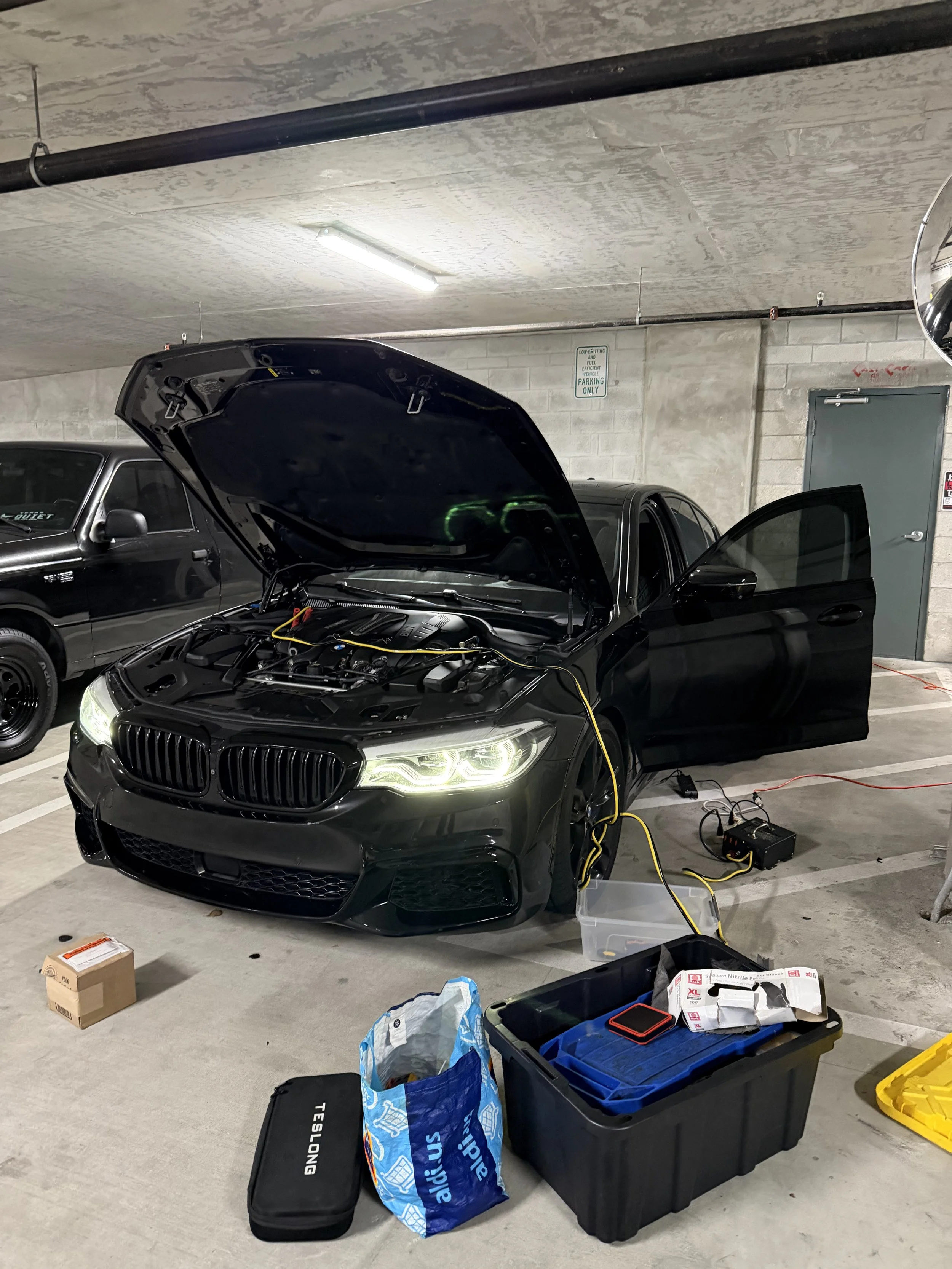 A black car with its hood open in a parking garage undergoing maintenance, surrounded by various tools, equipment, and supplies.