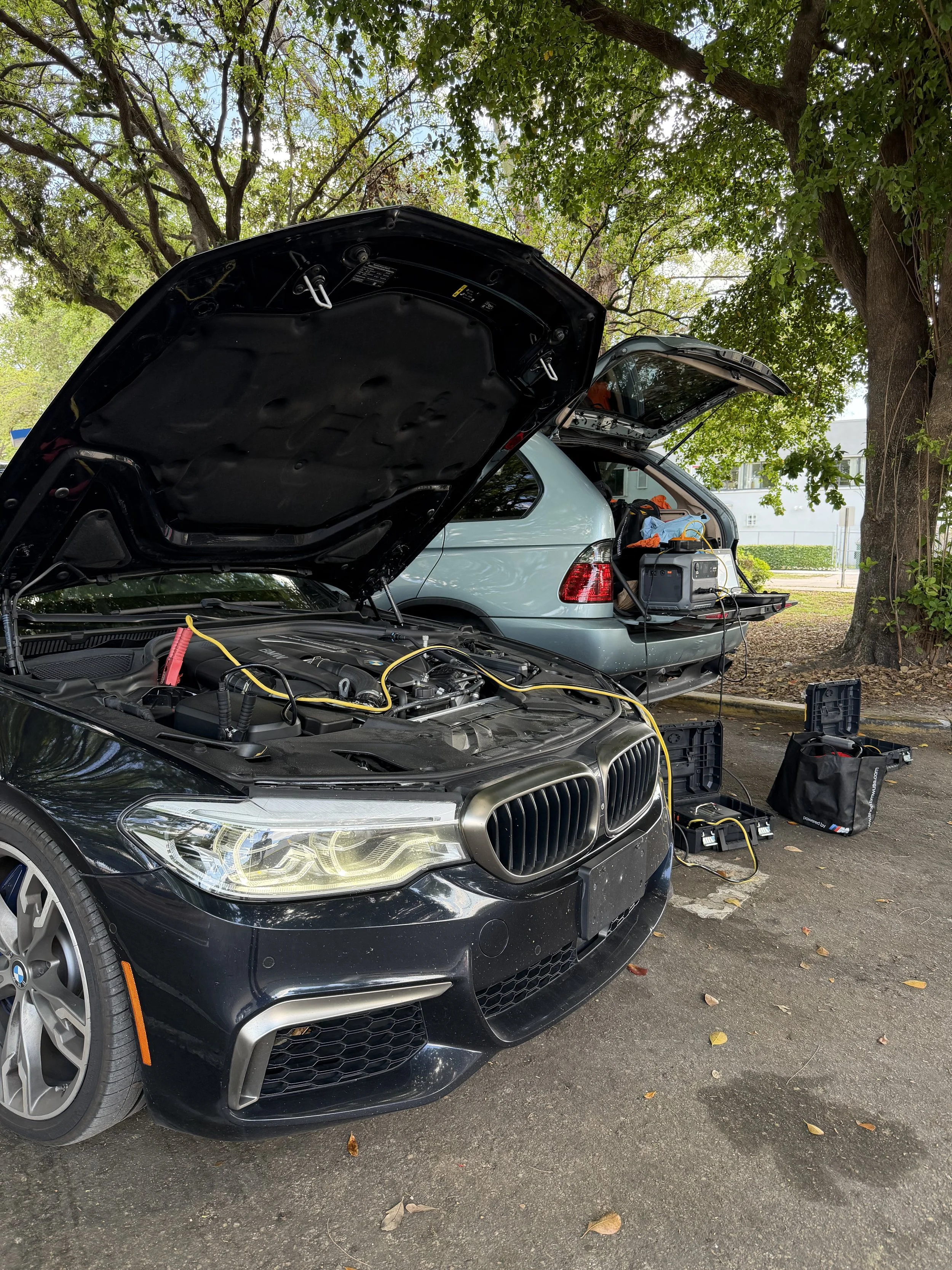 A black BMW car with its hood open, and a silver SUV with its rear hatch open, being serviced outdoors in a parking lot with trees and a building in the background.