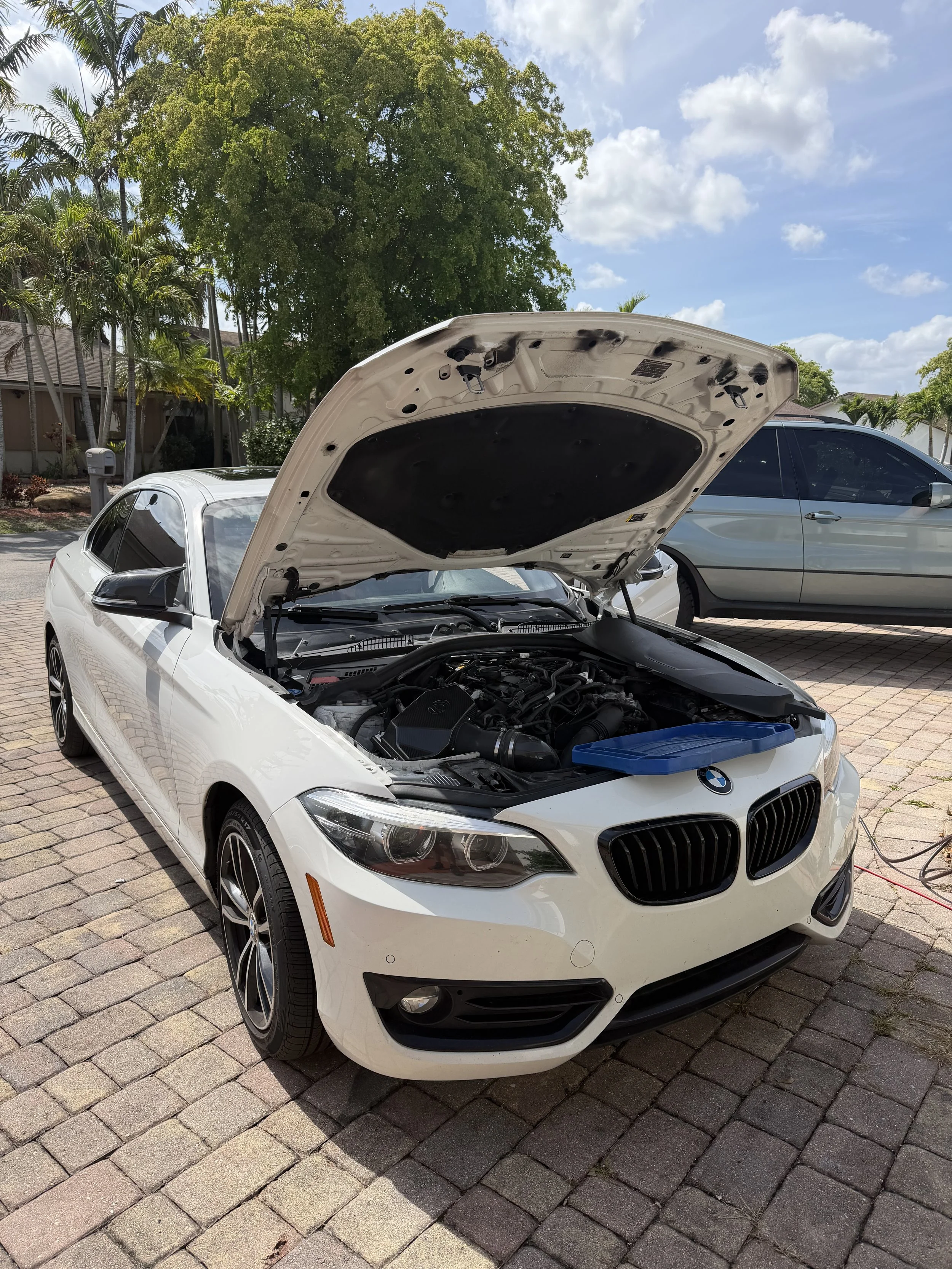 A white BMW with its hood open parked on a brick driveway in a residential neighborhood with trees and other parked cars visible in the background.