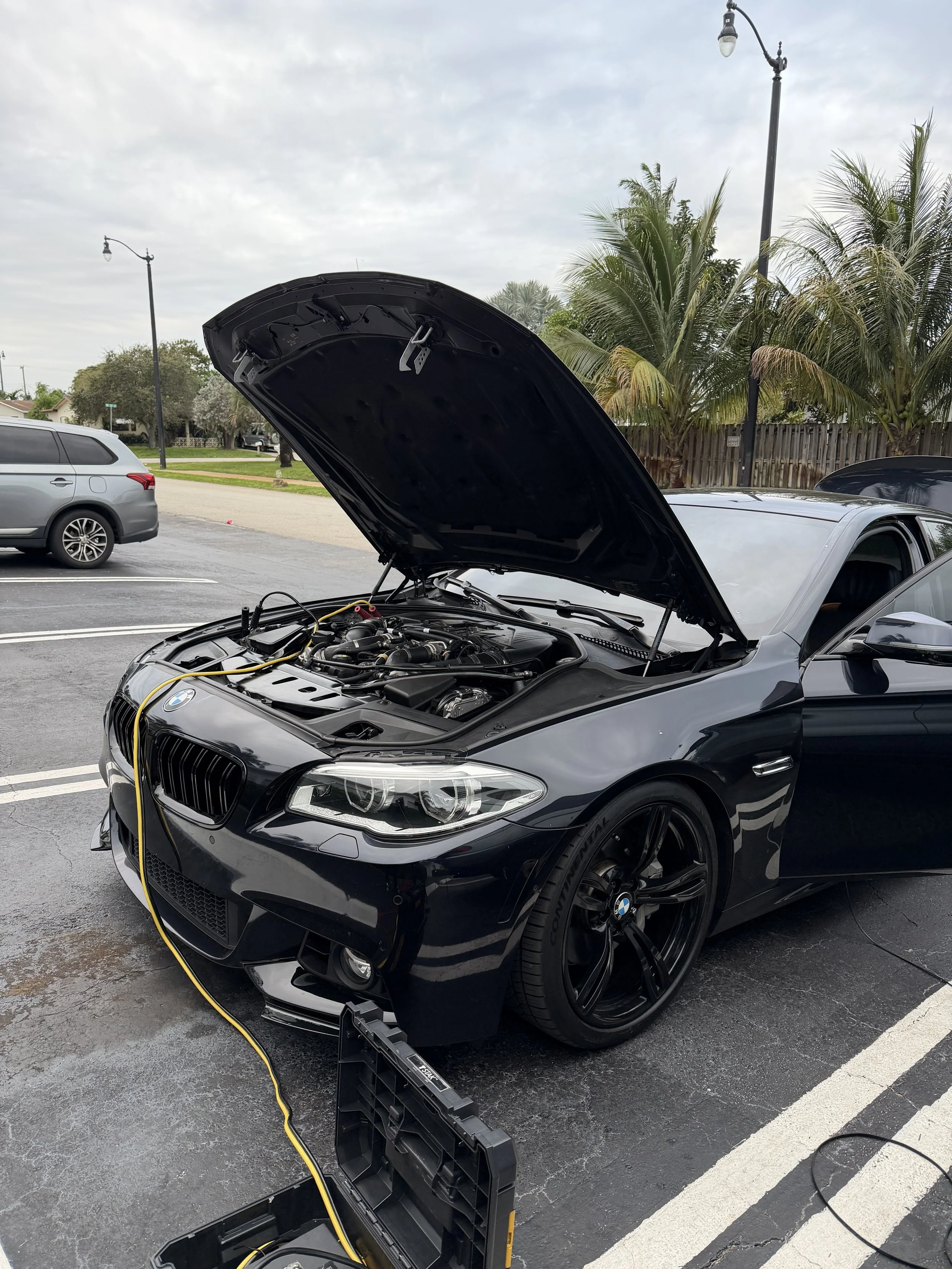 Black BMW with its hood open, placed in a parking lot during daytime, with a yellow jumper cable connected to the engine, and a battery pack on the ground.