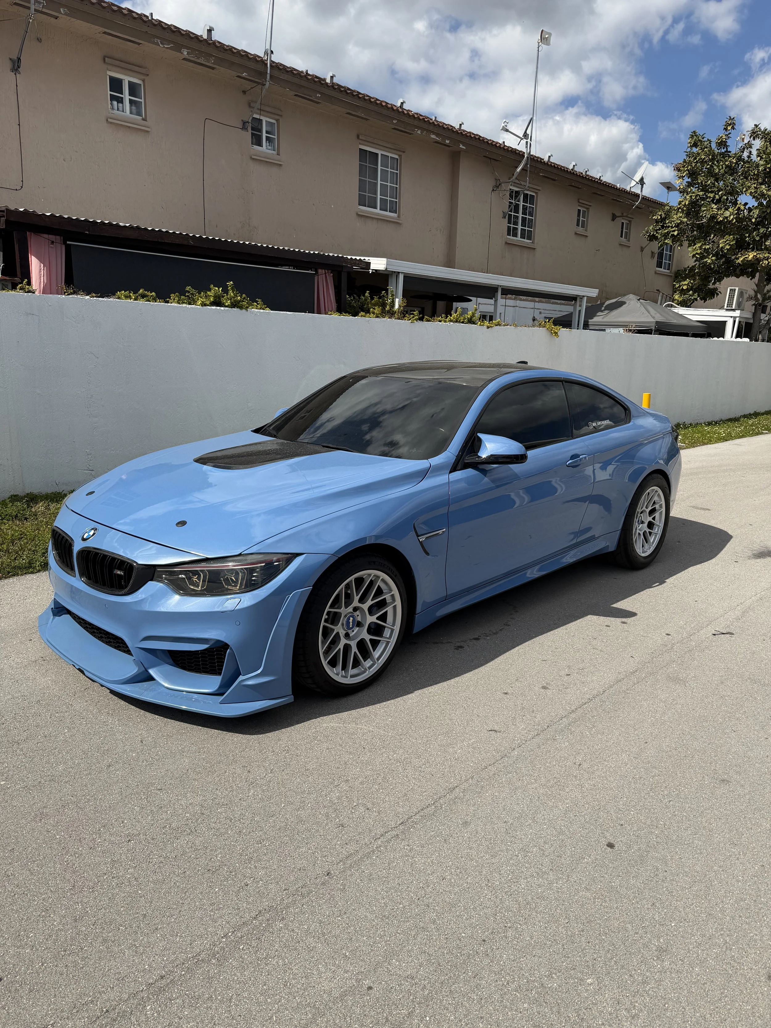 A blue BMW sports car parked on a concrete road with a white wall and beige residential building with multiple small windows and rooftop antennas in the background under a partly cloudy sky.