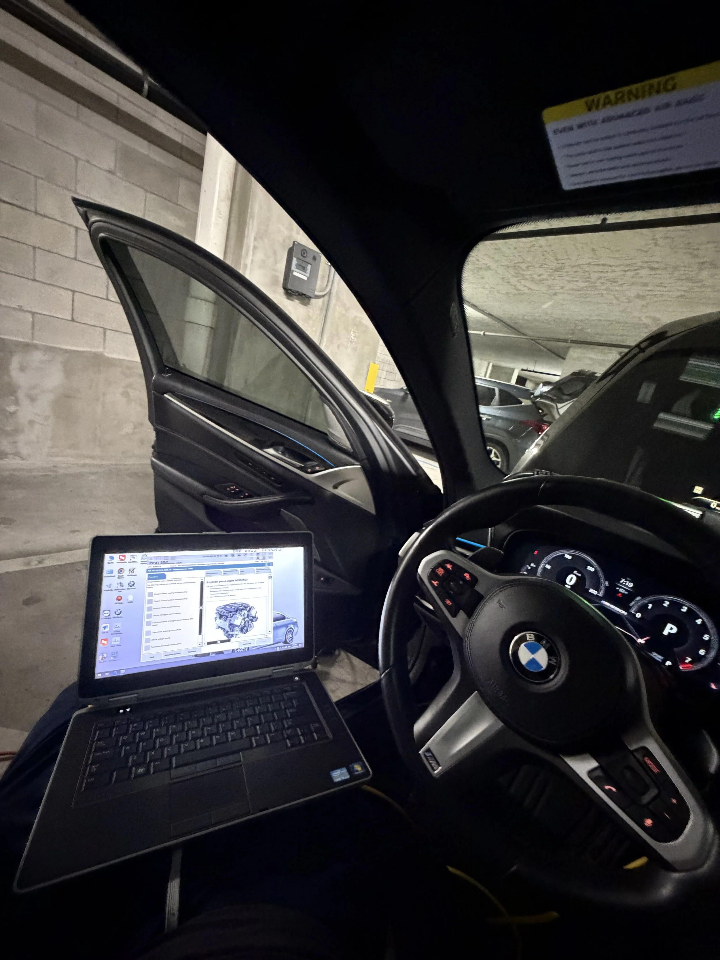 Inside a car in a parking garage, with a laptop on the driver's seat and various parked cars visible outside.