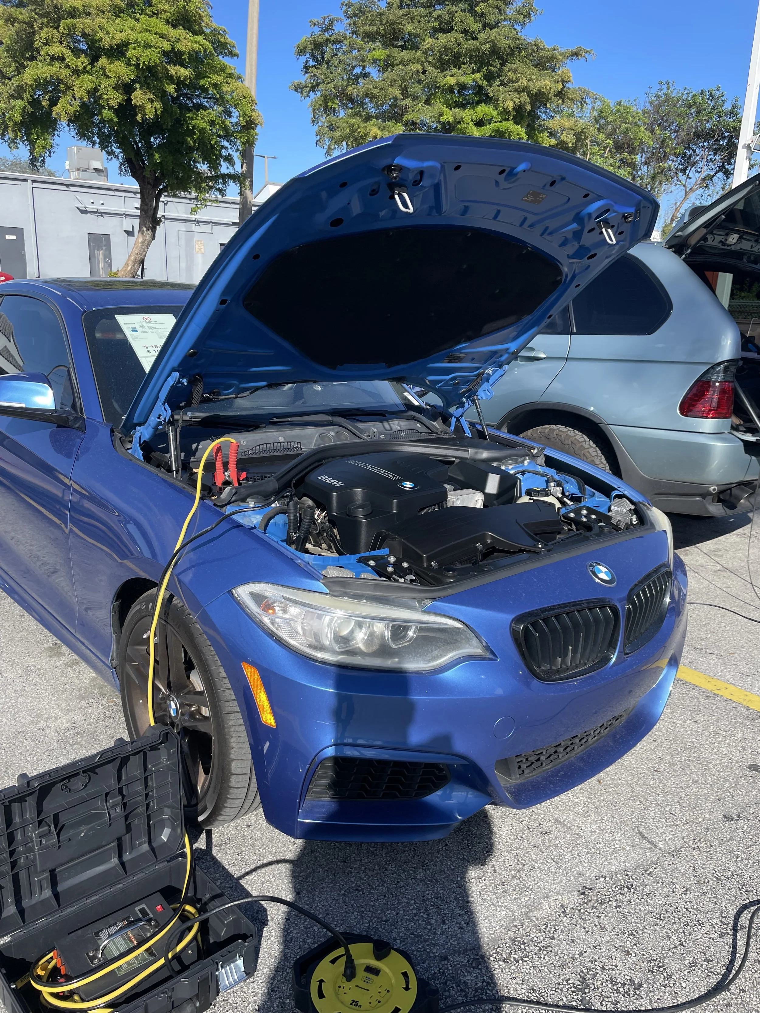 A blue BMW with its hood open being charged in a parking lot. There is a charging station connected to the car, with a yellow and black power supply on the ground. Trees and other vehicles are visible in the background.