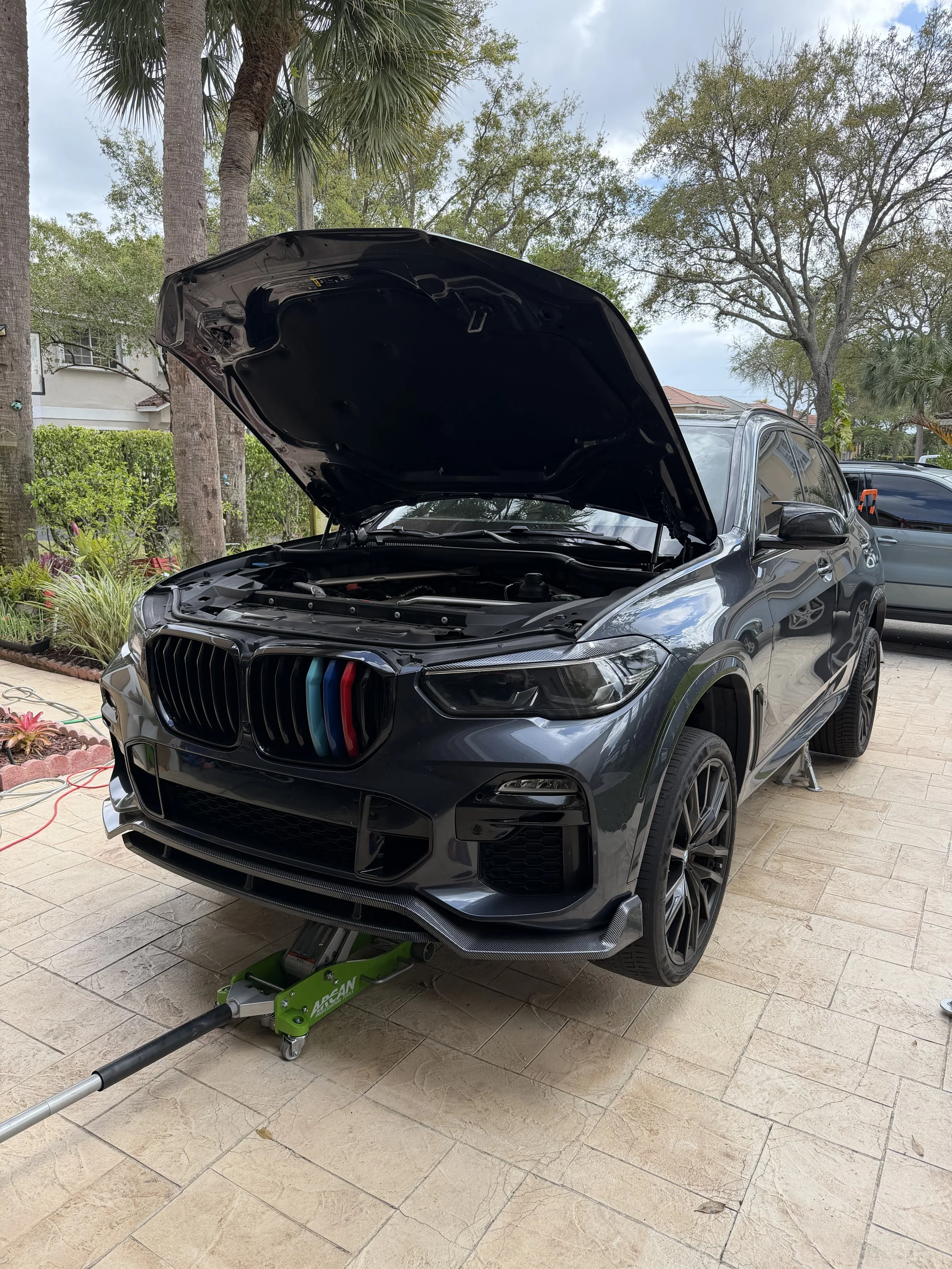 A black luxury SUV with its hood open, parked on a paved surface, being lifted by a green hydraulic car jack, outdoors with trees and a cloudy sky in the background.