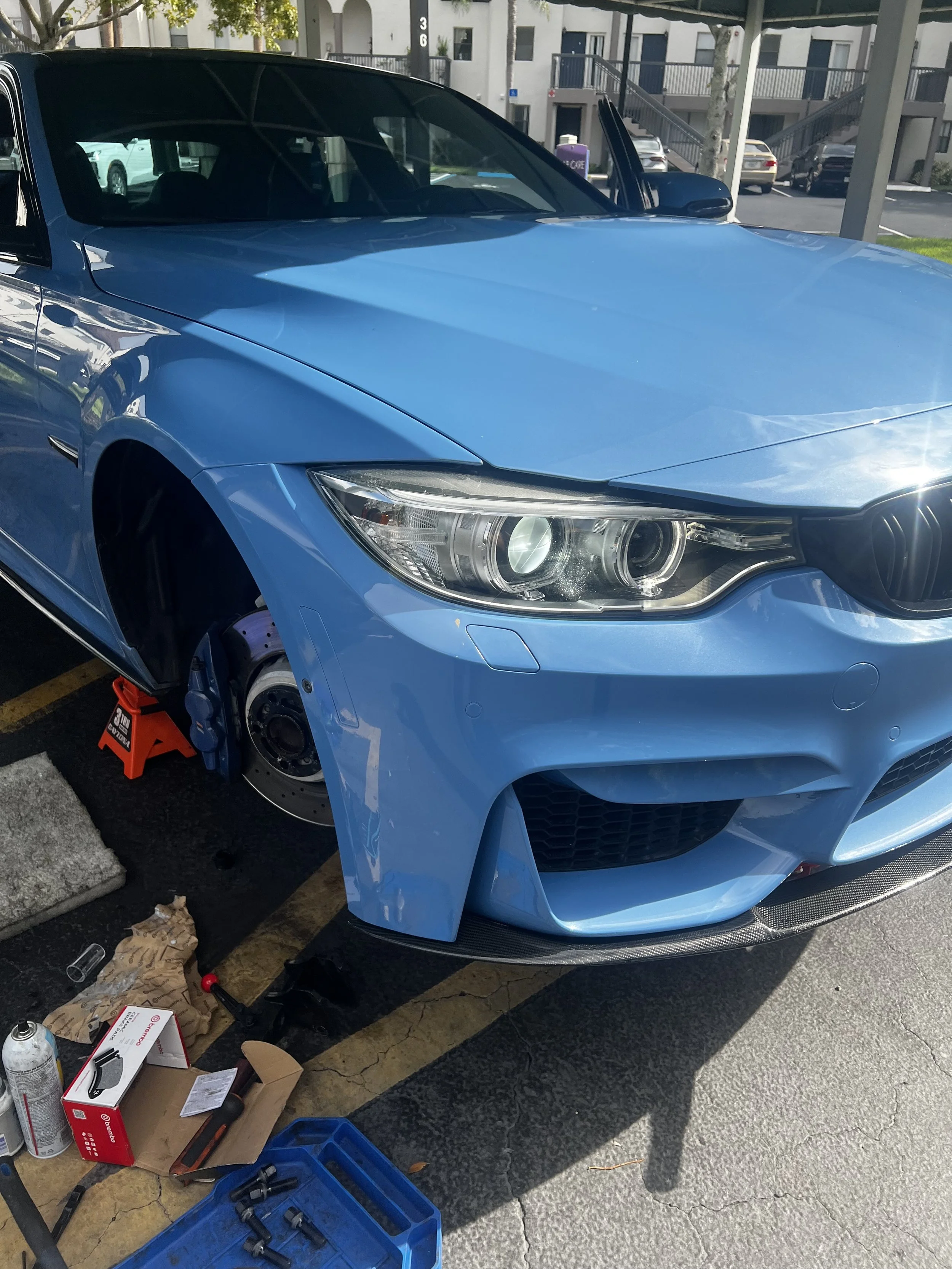 Blue car parked in a parking space with the front left wheel removed, placed on a red jack stand. Car's front bumper, headlights, and side mirror are visible. Tools and parts are scattered on a cardboard surface and the ground nearby, including a blu