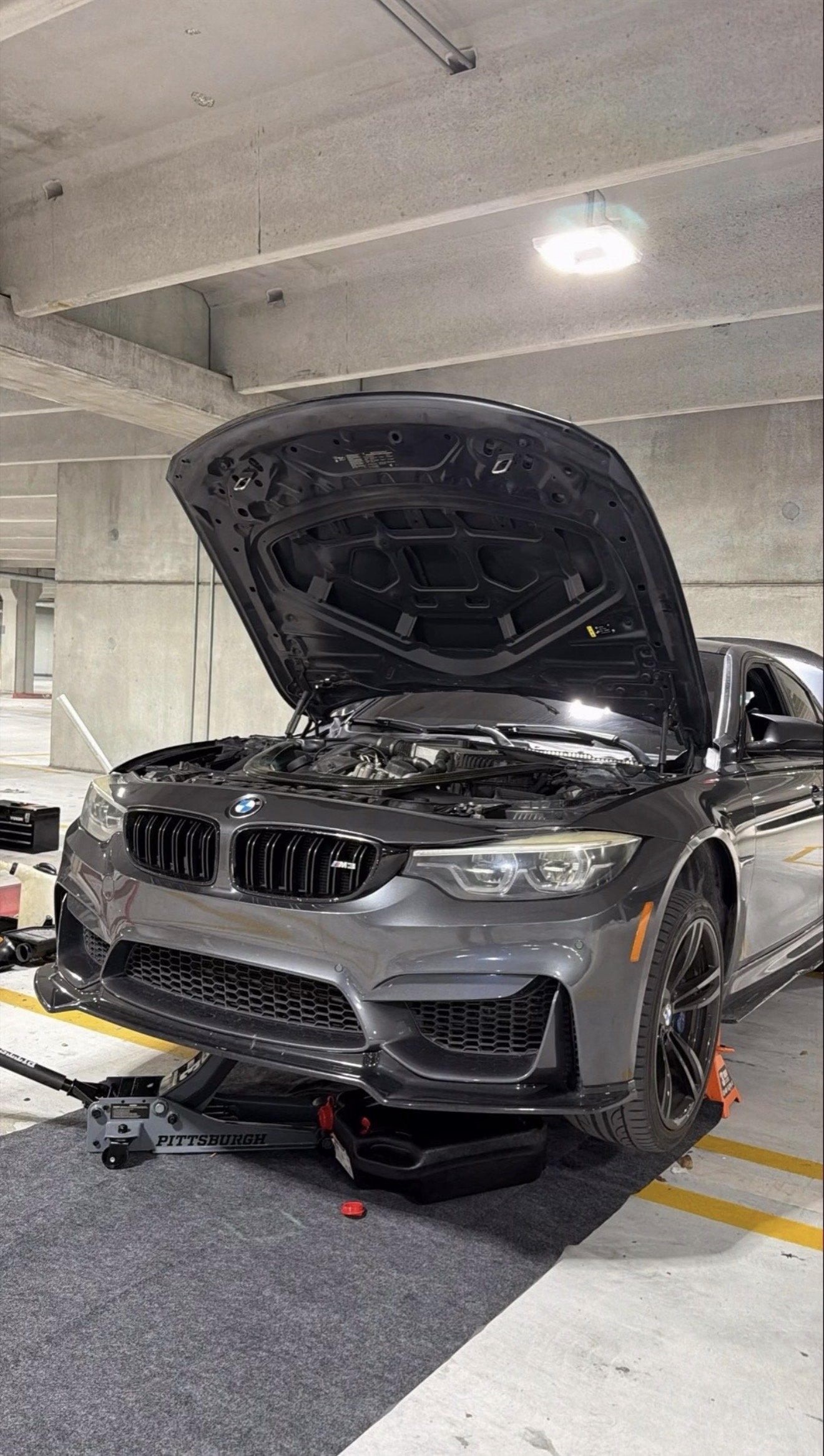 A gray BMW M4 with its hood open is parked in a concrete parking garage. It is on a lift or jack, with a tool chest nearby.