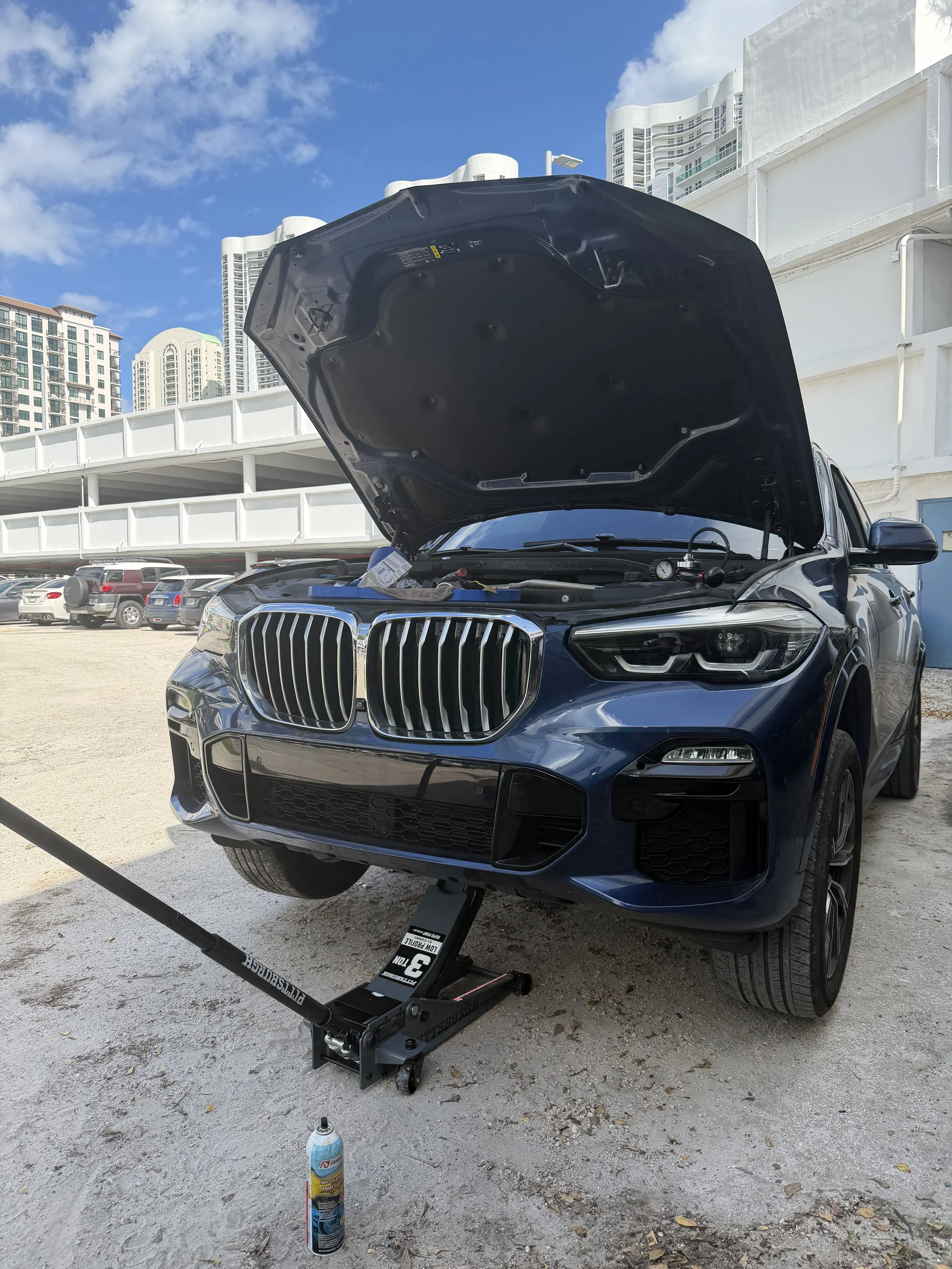 A black luxury SUV with its hood open, raised on a hydraulic lift in an outdoor parking lot. There is a spray can on the ground, with several other vehicles and tall buildings in the background under a partly cloudy sky.