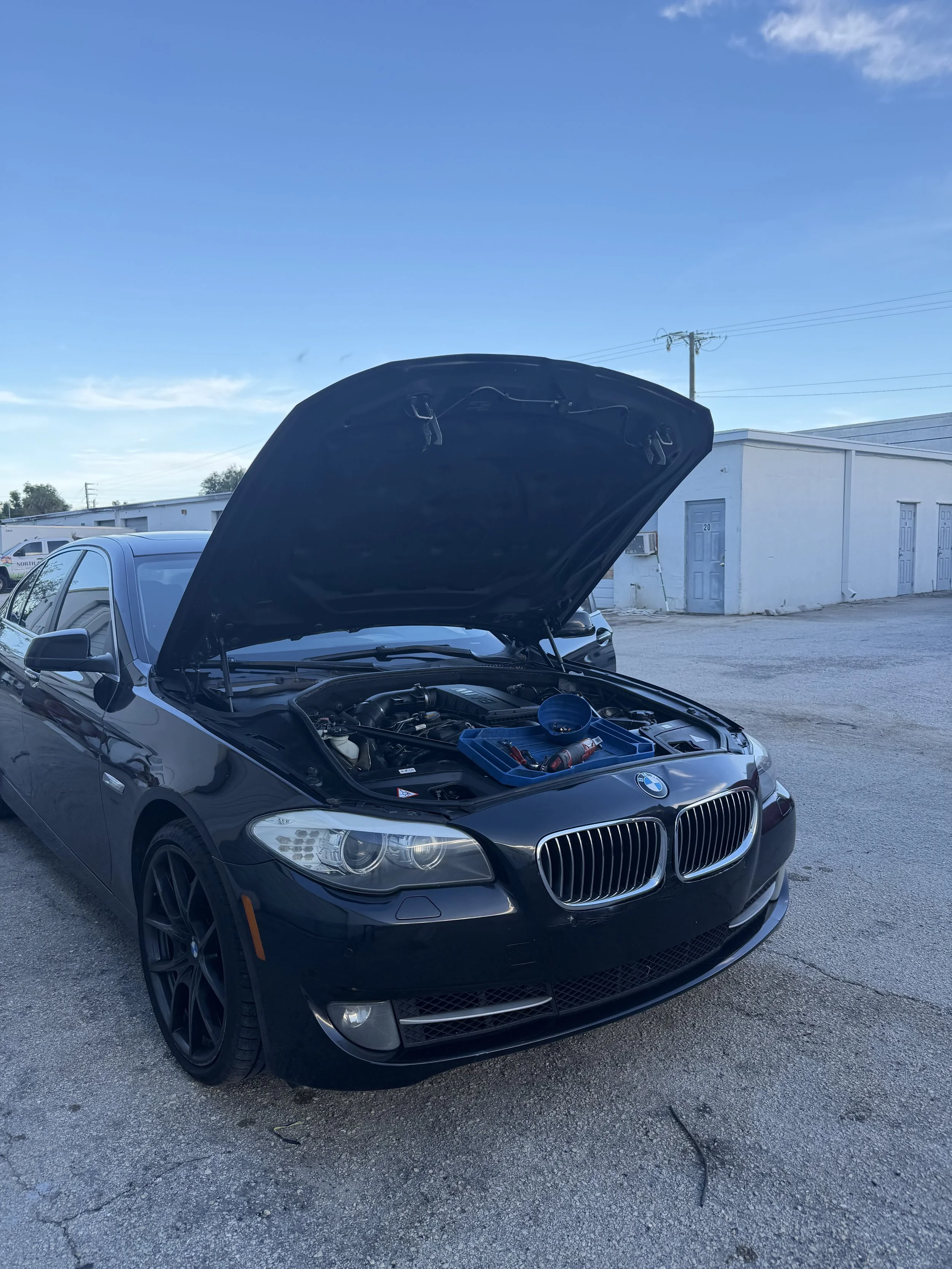 Black BMW car with hood open, parked outdoors near a white building and power lines, under a blue sky with some clouds.