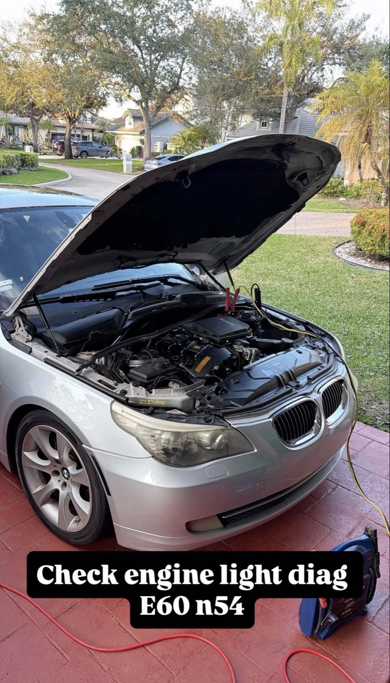 A white BMW with its hood open, revealing the engine. There is a diagnostic tool connected to the vehicle. The background shows a suburban neighborhood with houses and trees. Text overlays the image, reading 'Check engine light diag E60 n54'.