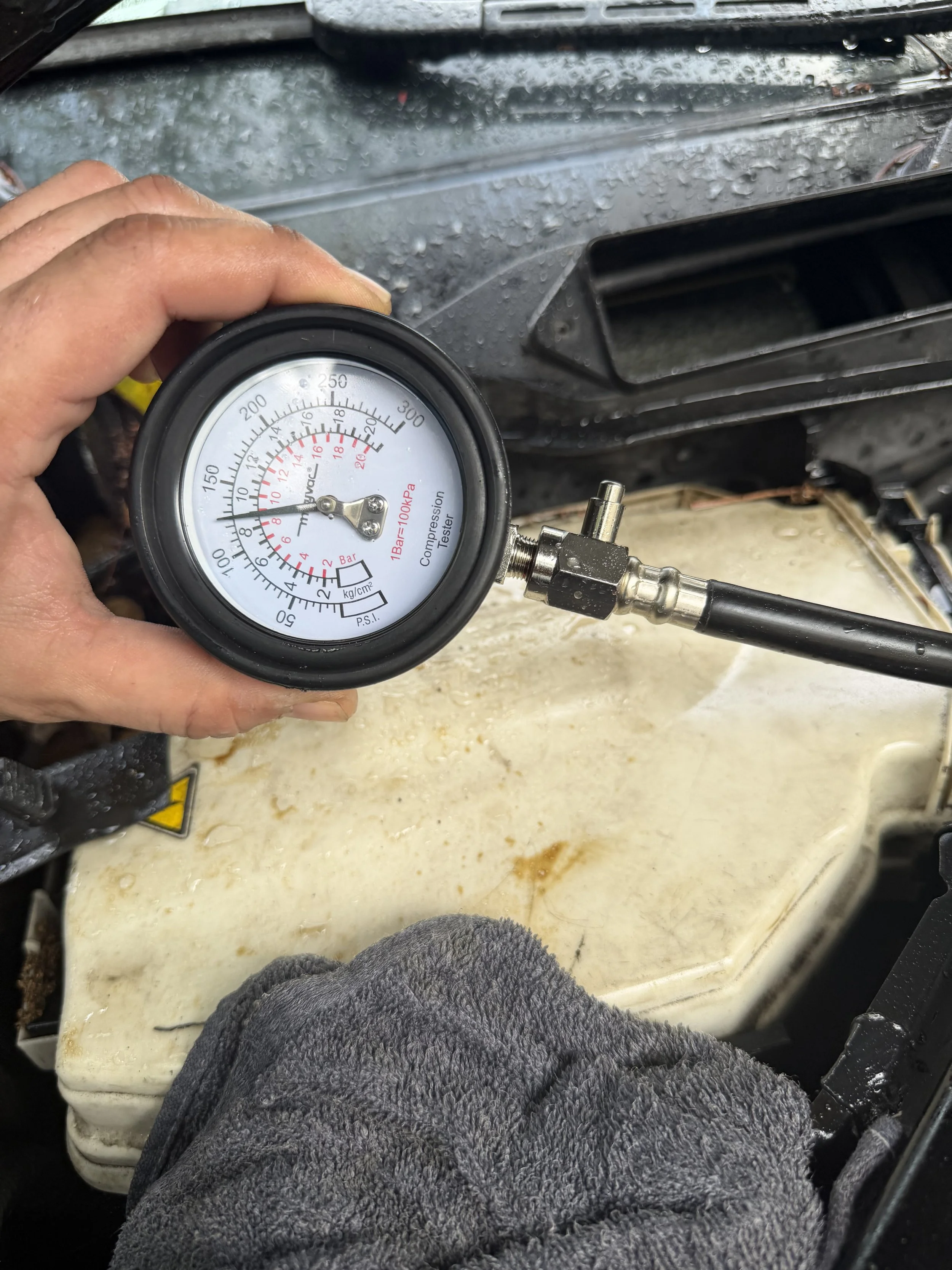 A person holding a tire pressure gauge in front of a car engine, with rain droplets visible on the windshield.