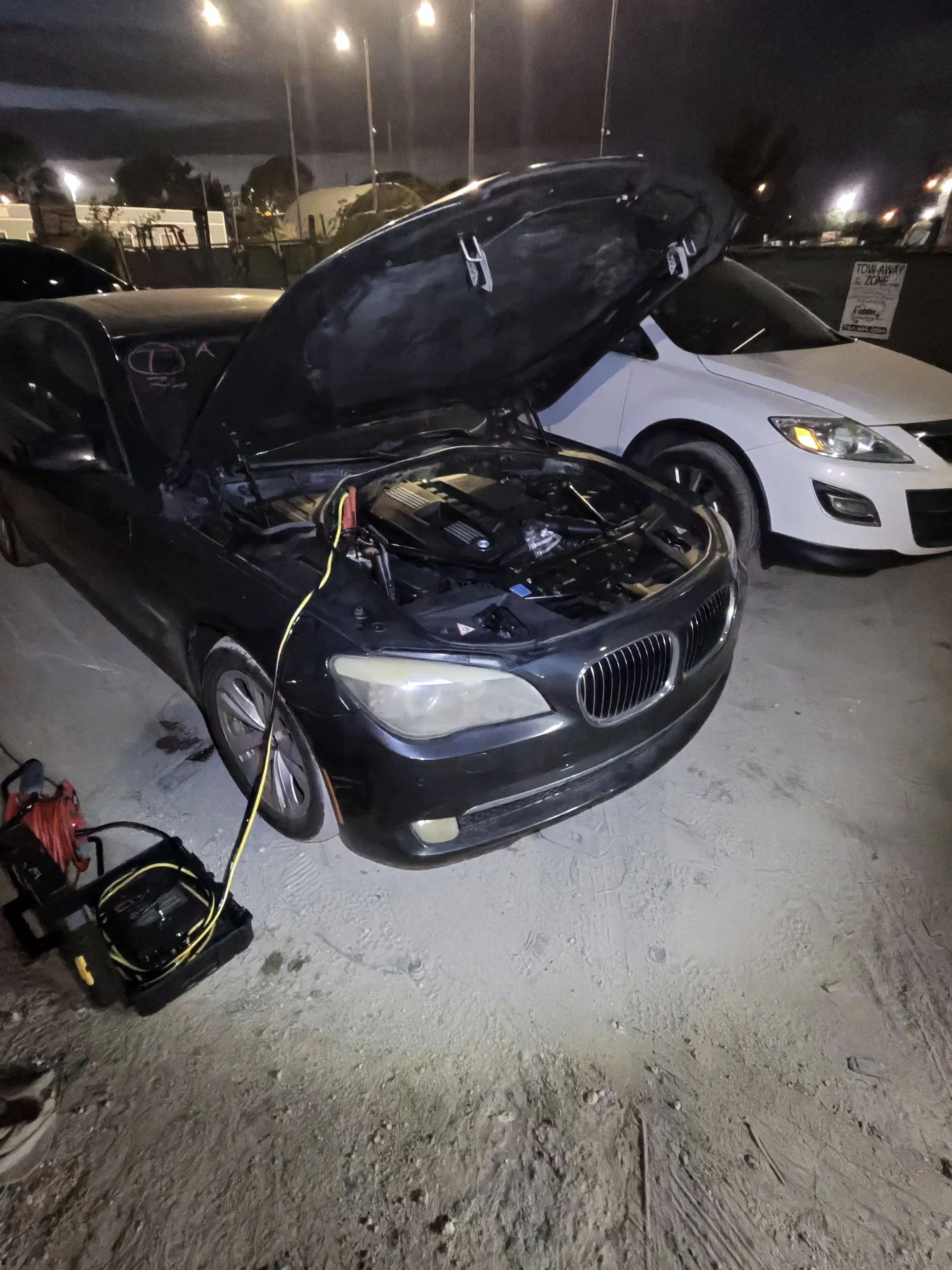 A black BMW car with its hood open, parked next to a white car at night, with electrical equipment and cables on the ground nearby.