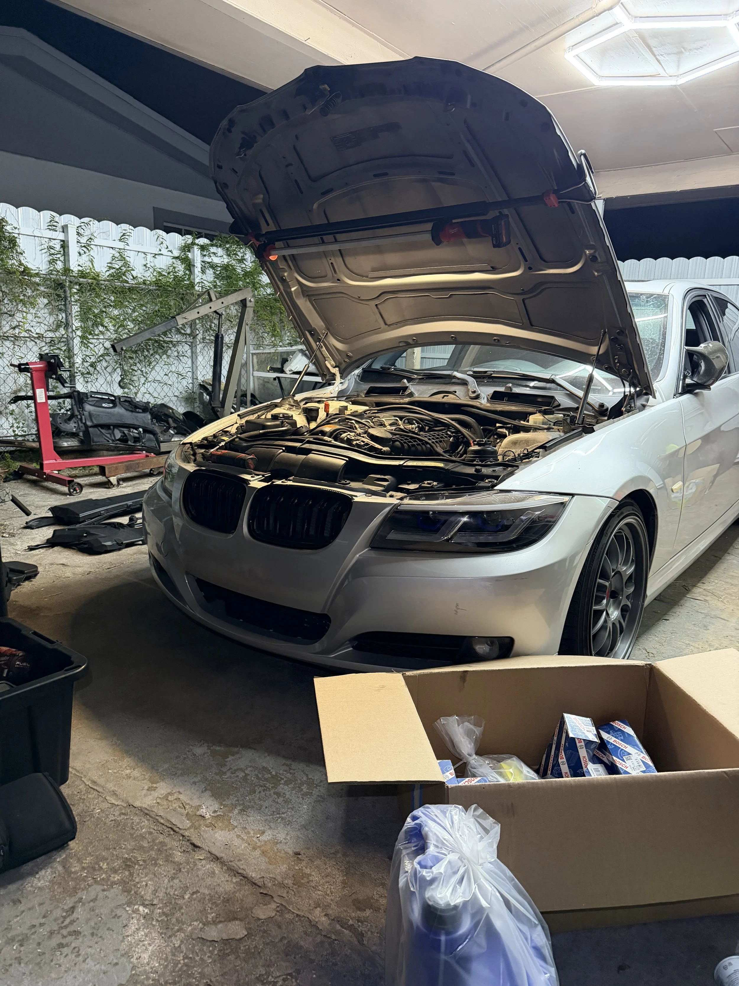 A silver car with its hood open in a garage or workshop, surrounded by various tools, parts, and cardboard boxes, indicating ongoing vehicle repairs or maintenance.
